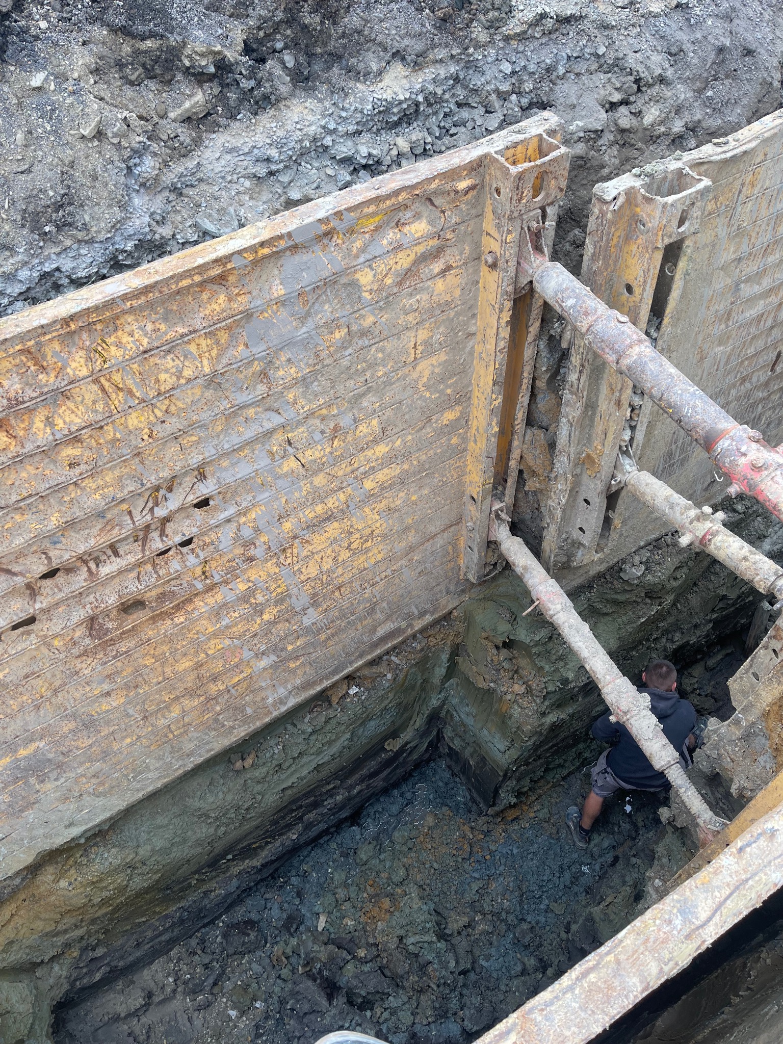 Głęboki wykop zabezpieczony metalowymi szalunkami, widoczny pracownik na dnie, warstwy ziemi o różnej strukturze i kolorze.