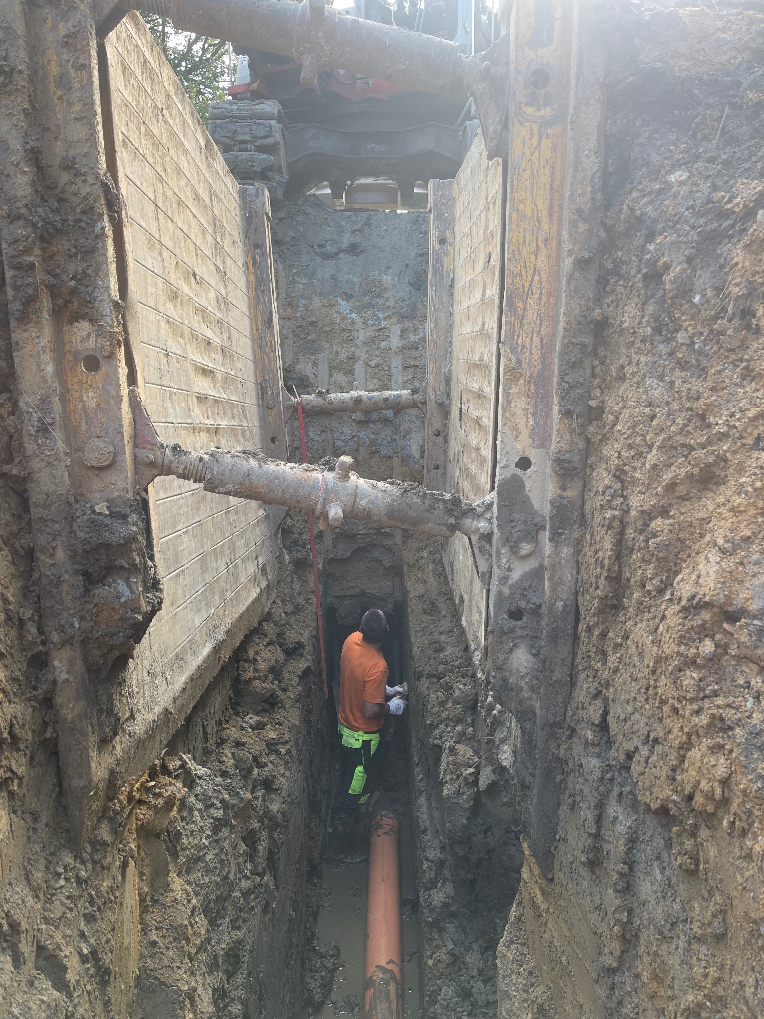 Głęboki wykop zabezpieczony stalowymi szalunkami, wewnątrz pracownik w odblaskowej odzieży łączy pomarańczową rurę; na górze widoczny fragment koparki.