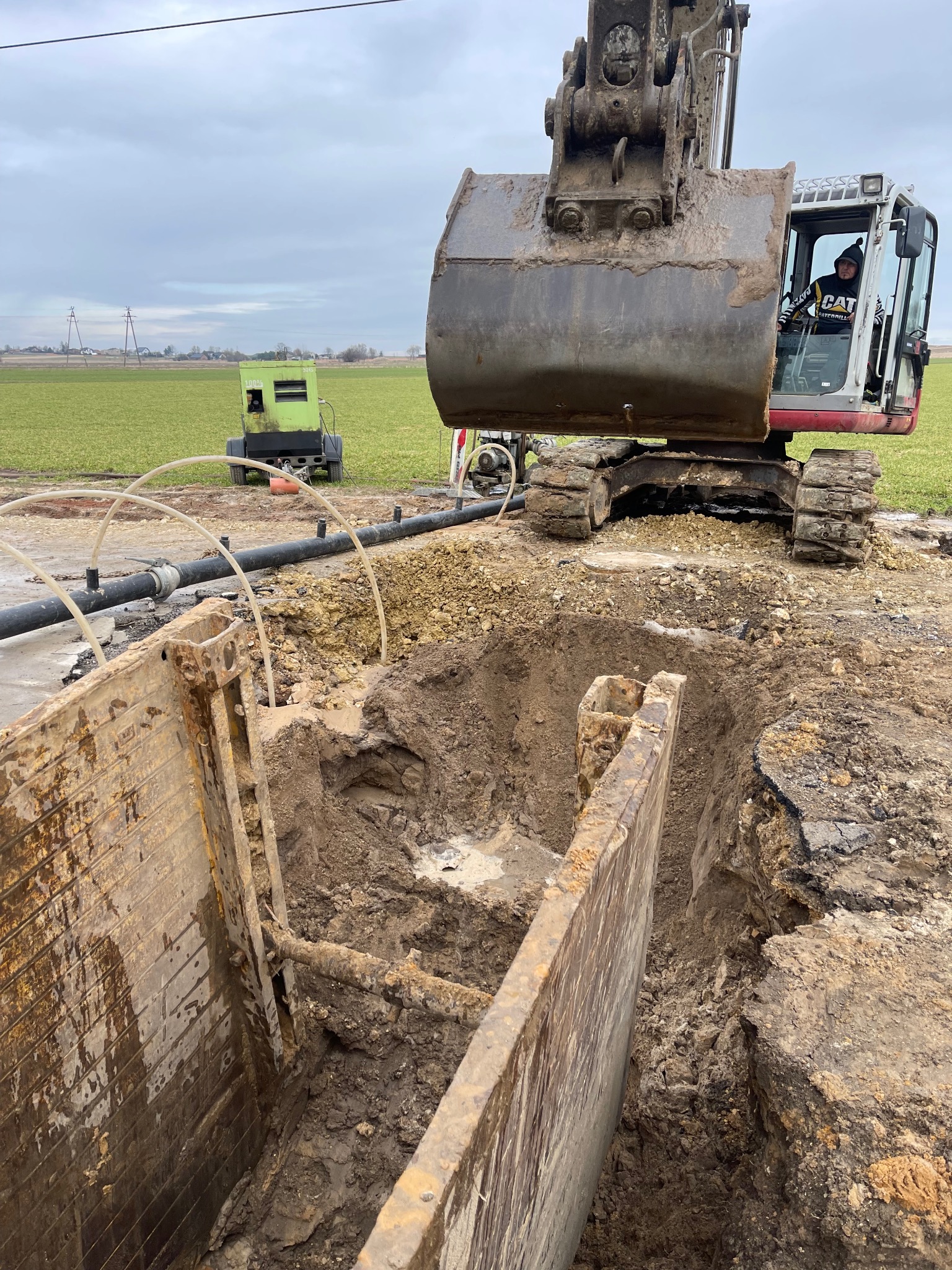 Operator koparki Caterpillar wykonuje wykop zabezpieczony stalową szalunką, widoczne rury i agregat prądotwórczy na tle zielonego pola pod pochmurnym niebem.
