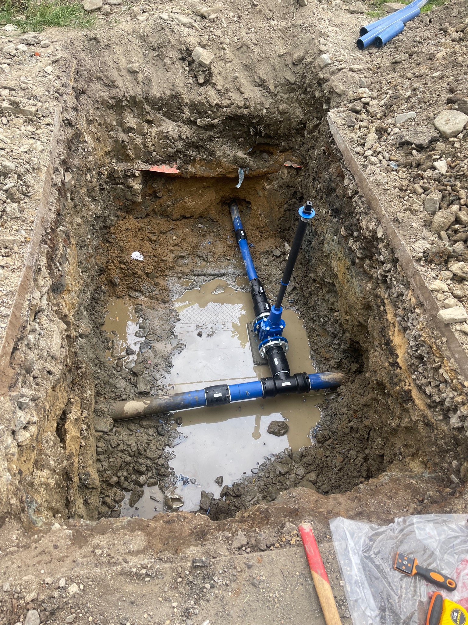 Wykop z ułożonymi rurami wodociągowymi i kanalizacyjnymi wypełniony wodą, widoczne narzędzia pracy na brzegu wykopu.