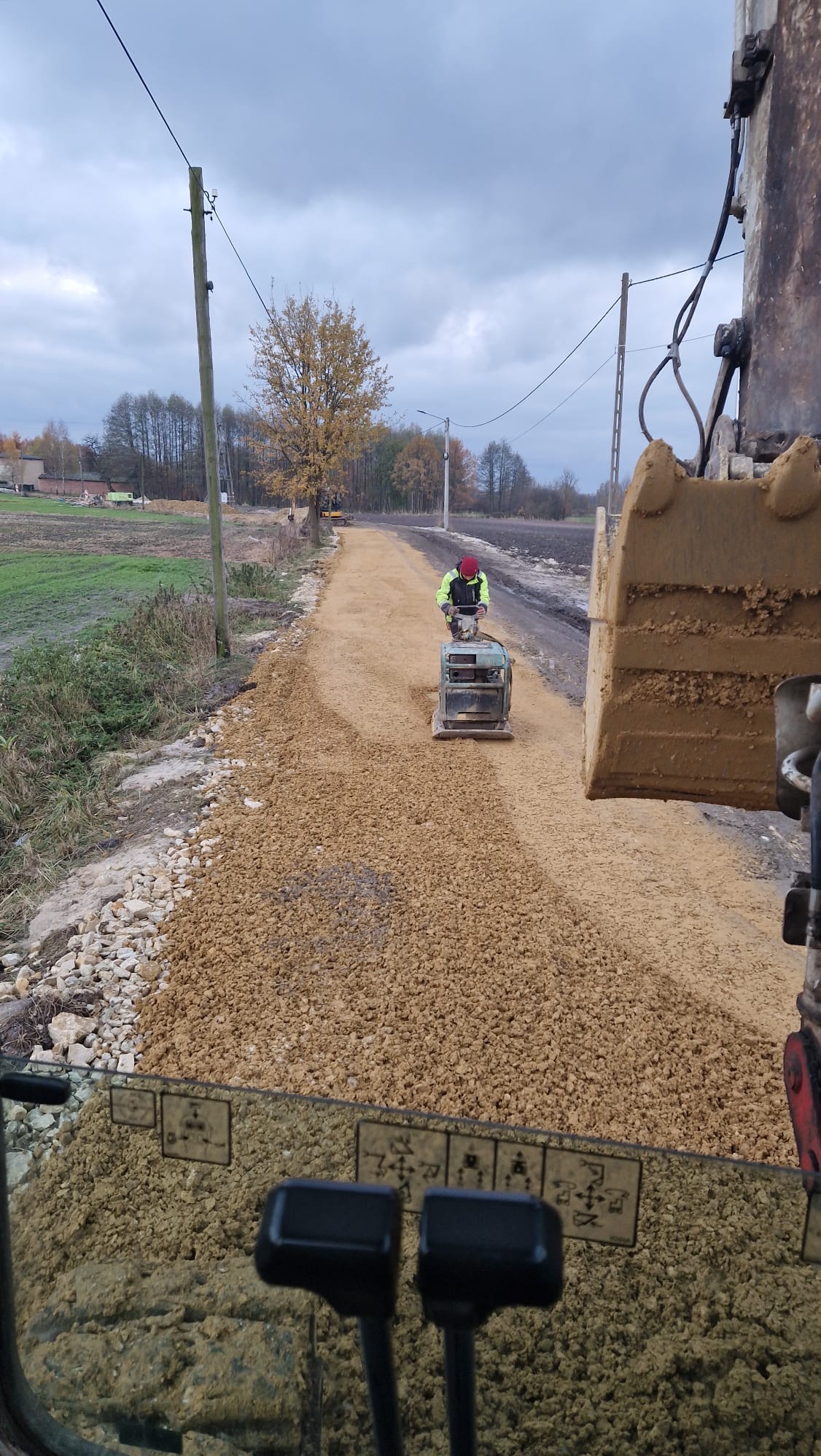 Operator w kamizelce odblaskowej zagęszcza nową nawierzchnię drogi ubijarką wibracyjną, widok z kabiny koparki z widocznymi dźwigniami sterującymi i łyżką koparki w kadrze.