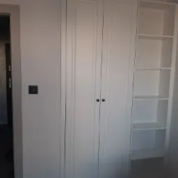 Biała szafa wnękowa z dwiema zamkniętymi częściami z czarnymi gałkami i otwartą półką z jasnymi półkami, widoczna obok włącznika światła i fragmentu futryny drzwiowej