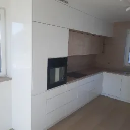 Nowoczesna, jasna kuchnia z białymi, lakierowanymi frontami i drewnianymi akcentami w Sławnie. Widoczna zabudowa z piekarnikiem i płytą indukcyjną oraz oświetlenie punktowe.