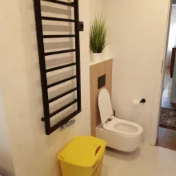 Nowoczesna łazienka z wiszącą toaletą, czarnym grzejnikiem drabinkowym, rośliną w doniczce i ż&oacute;łtym koszem na pranie, wykończona płytkami imitującymi marmur.