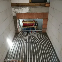 Rozdzielacz ogrzewania podłogowego z rurami ułożonymi na siatce w surowym pomieszczeniu z bloczków.