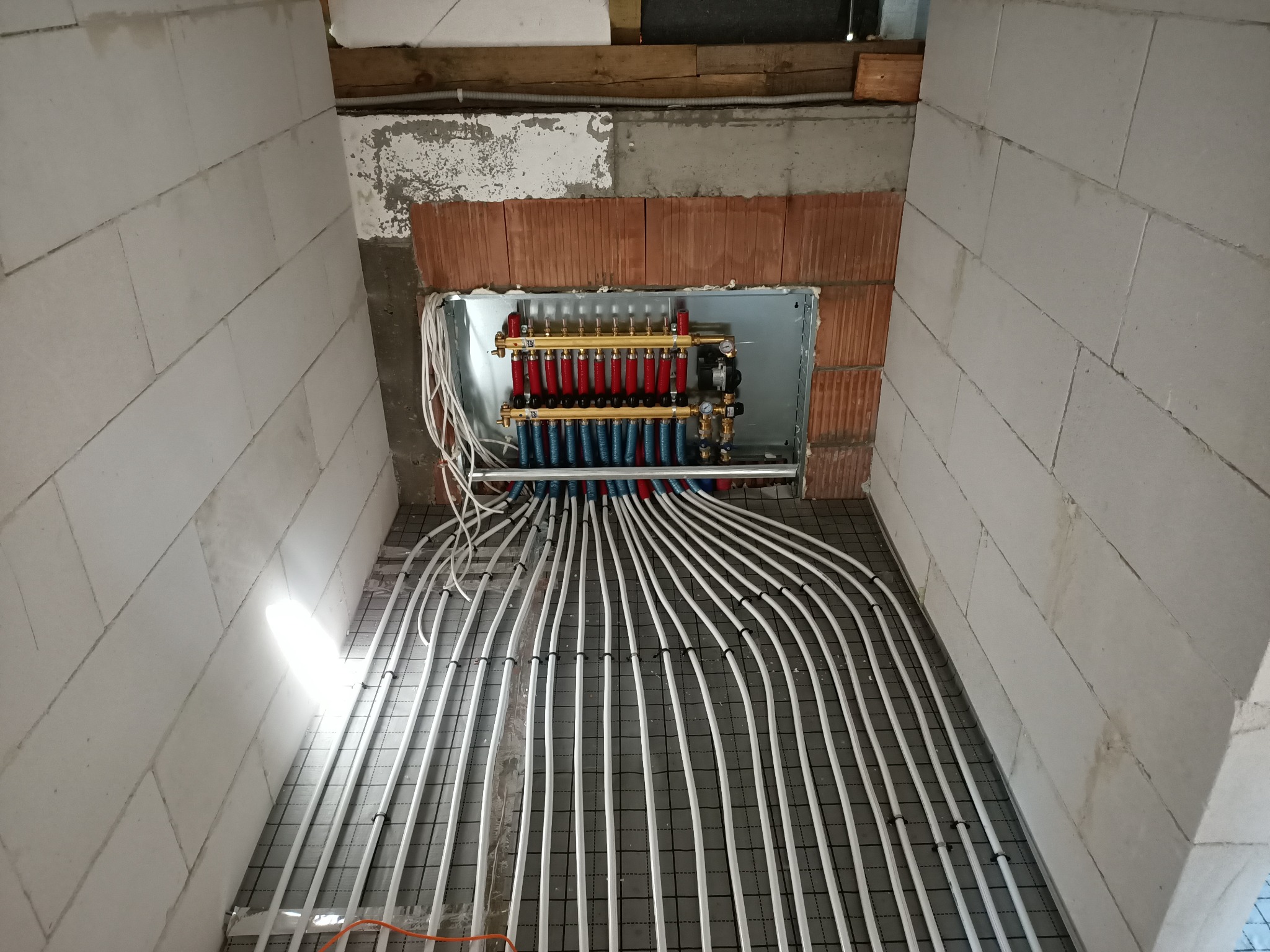 Rozdzielacz ogrzewania podłogowego z rurami ułożonymi na siatce w surowym pomieszczeniu z bloczków.