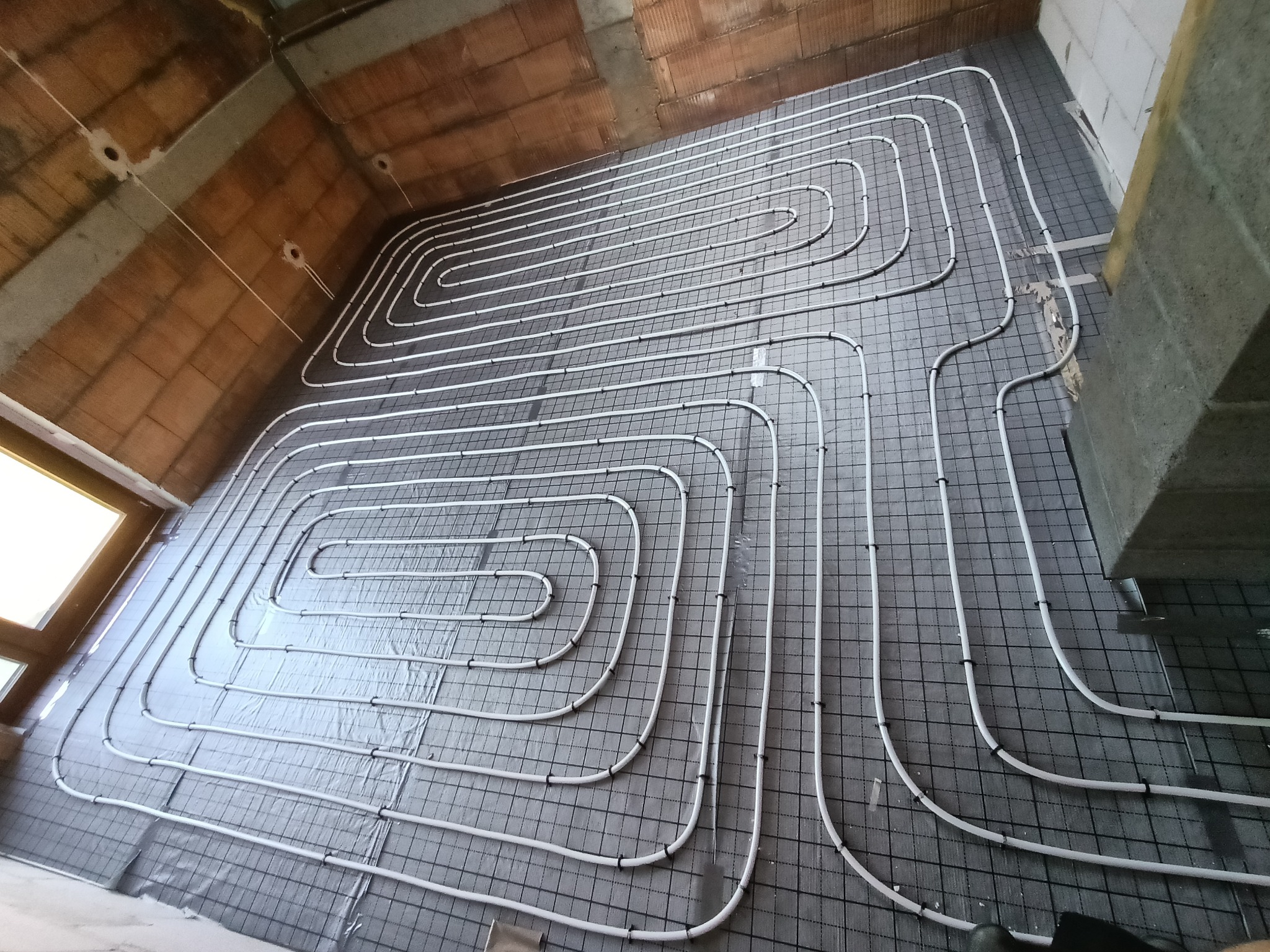 Białe rury ogrzewania podłogowego ułożone w pętle na czarnej siatce, widok z góry, ściany z cegły i betonu w surowym stanie.