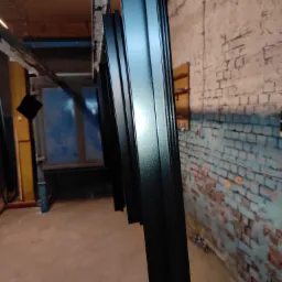 Czarne, metalowe profile zawieszone na wieszakach w hali produkcyjnej, przygotowane do dalszej obróbki lub po malowaniu, z fragmentem pieca w tle i ceglaną ścianą.