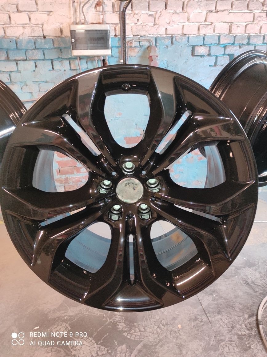 Błyszcząca, czarna felga samochodowa zawieszona na haku, z widocznym odbiciem światła i fragmentem ceglanej ściany w tle.