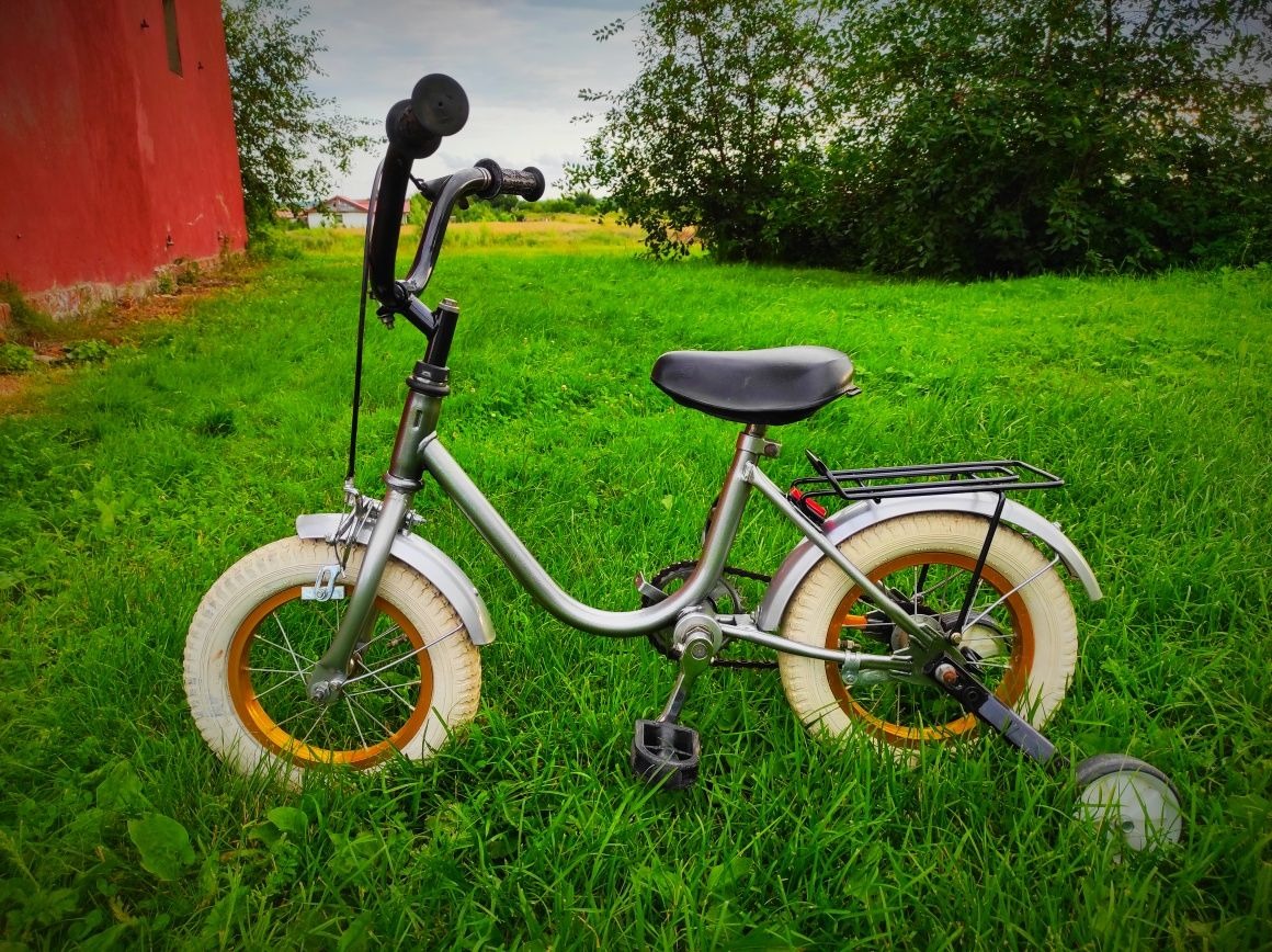 Dziecięcy rower w srebrnej ramie z białymi oponami i złotymi felgami, stojący na zielonej trawie, z czerwonym budynkiem w tle.