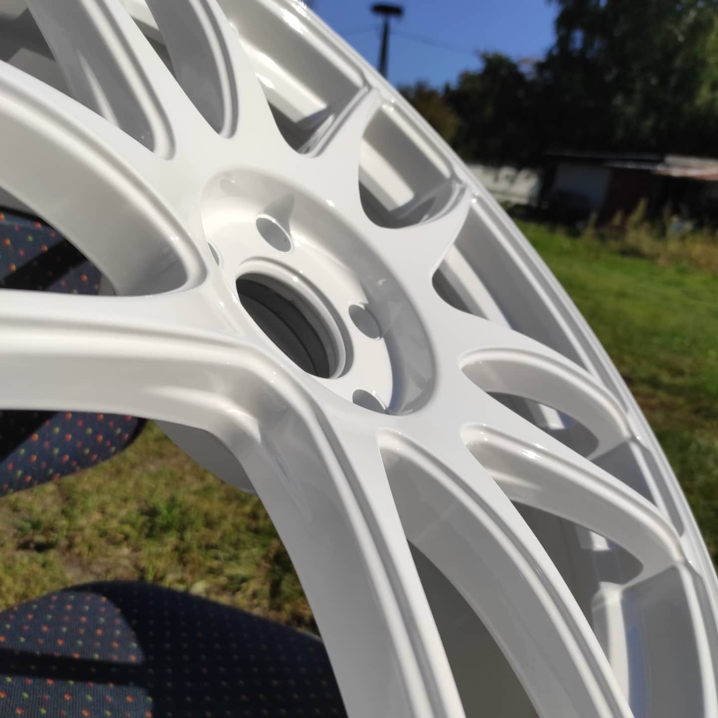 Biała felga samochodowa z lekkiego stopu, świeżo po renowacji, z połyskiem, sfotografowana w plenerze na tle zielonej trawy i błękitnego nieba.