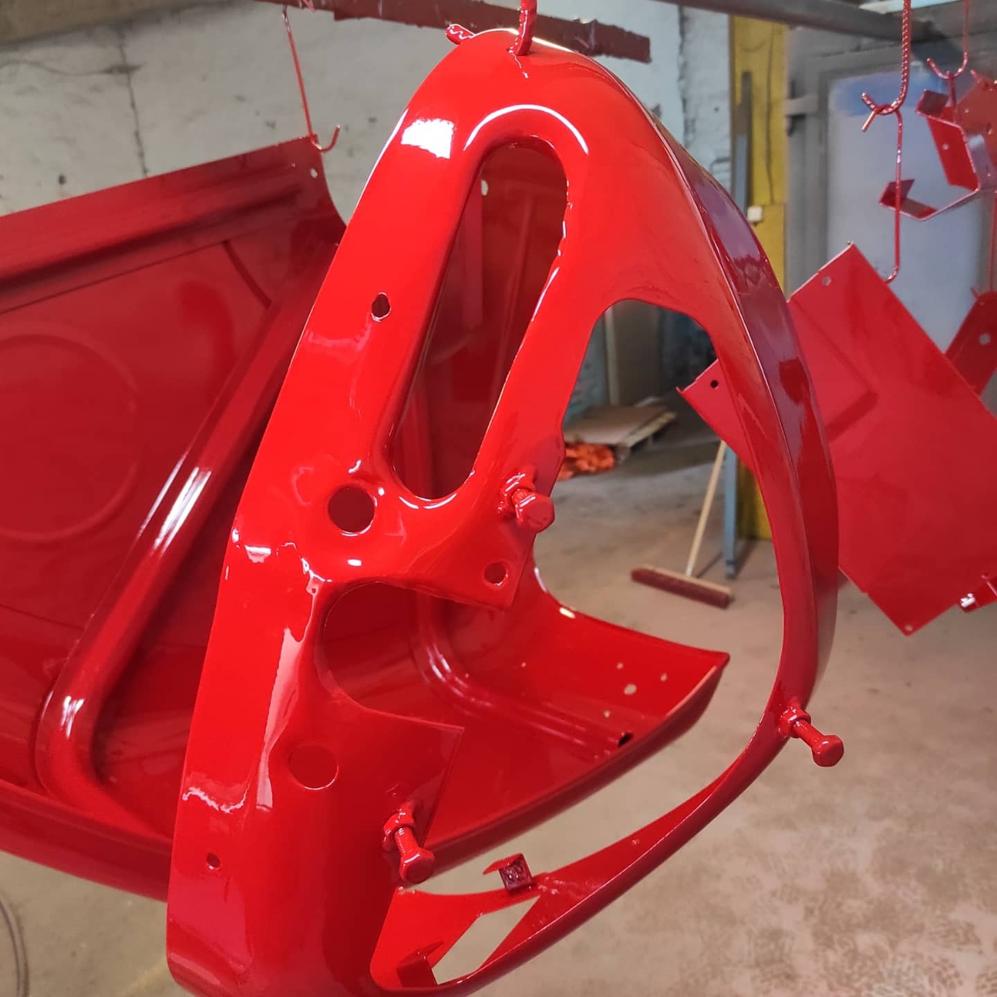 Czerwone elementy karoserii samochodowej, świeżo po malowaniu proszkowym, zawieszone na metalowym stelażu w warsztacie. Widoczny połysk farby i detale mocowań.