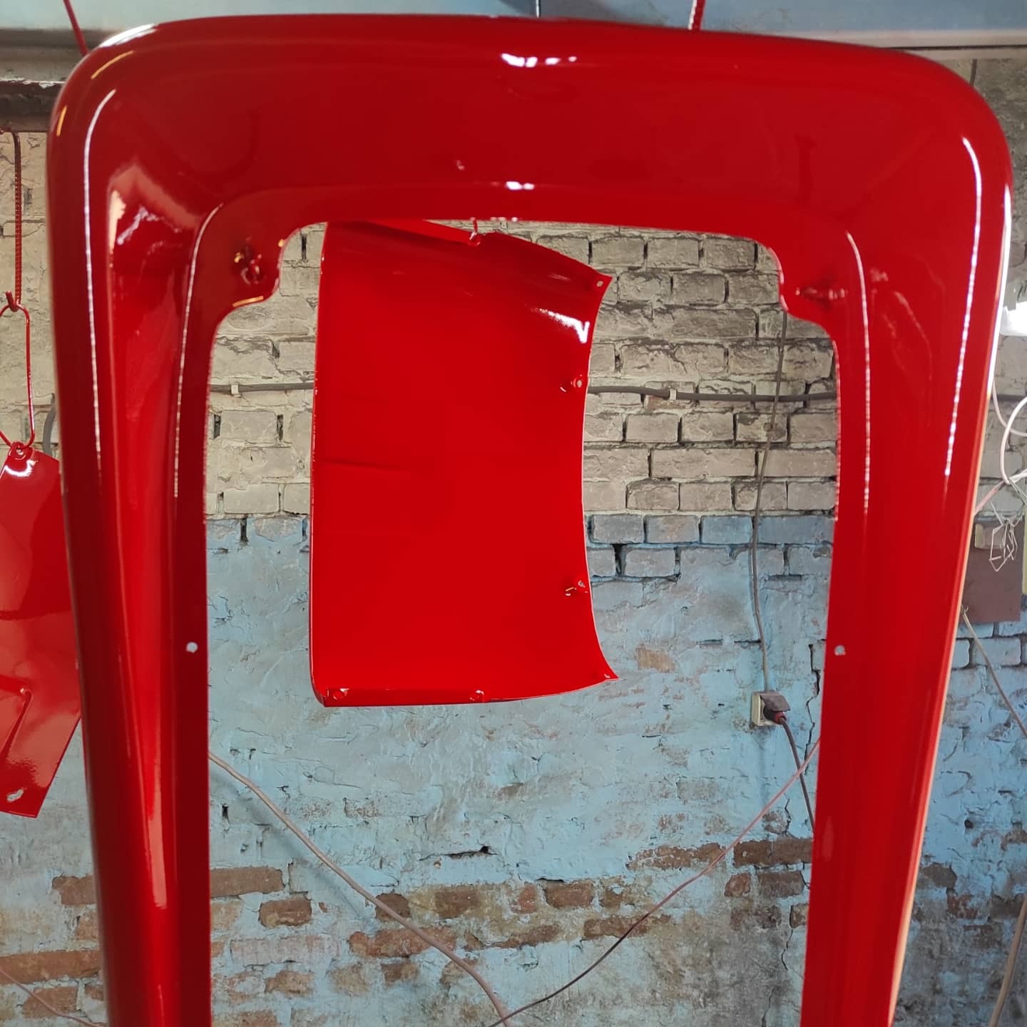 Elementy metalowe po malowaniu proszkowym w intensywnym czerwonym kolorze, zawieszone na hakach w warsztacie, z widoczną ścianą z cegieł w tle.