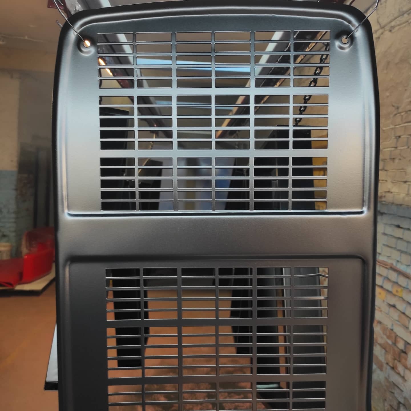 Metalowa obudowa z perforacją, świeżo po nałożeniu szarej powłoki, zawieszona na haku w warsztacie, z widoczną ceglaną ścianą w tle.