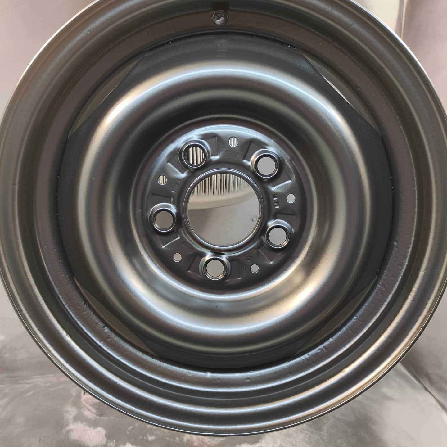 Metalowa felga samochodowa po renowacji, pokryta świeżą, srebrną farbą, widoczne detale mocowań i otwory wentylacyjne.