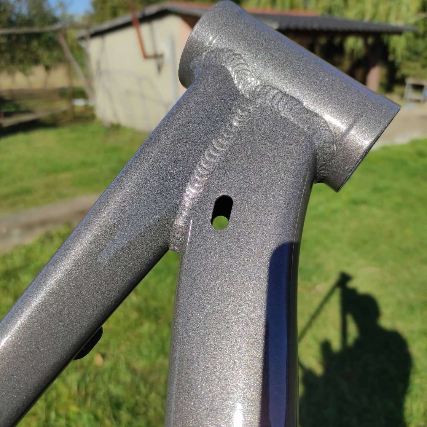 Metalowa rama rowerowa po obróbce, z widocznymi śladami spawania i świeżą, szarą powłoką. W tle zieleń trawy i zarys budynku.