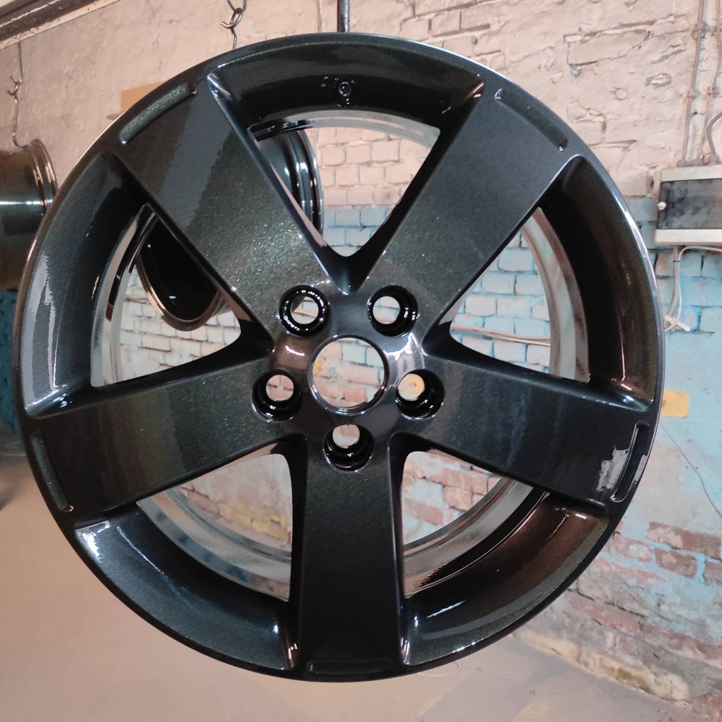 Pomalowana na ciemny grafit felga aluminiowa zawieszona na haku w warsztacie, widoczne ślady po niedokładnym oczyszczeniu powierzchni przed malowaniem.