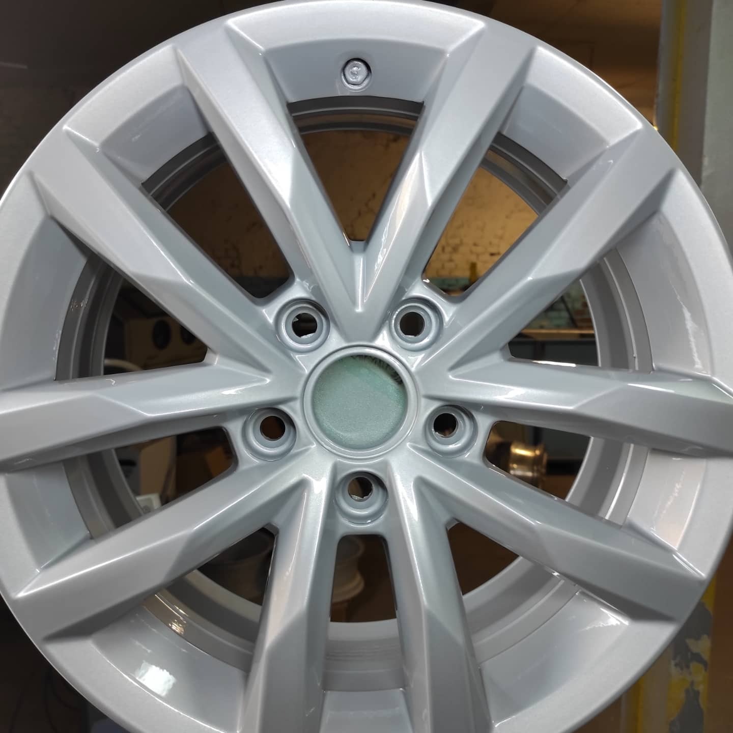 Aluminiowa felga samochodowa po renowacji, świeżo pomalowana na jednolity, srebrny kolor, widoczna w zbliżeniu na tle warsztatu.