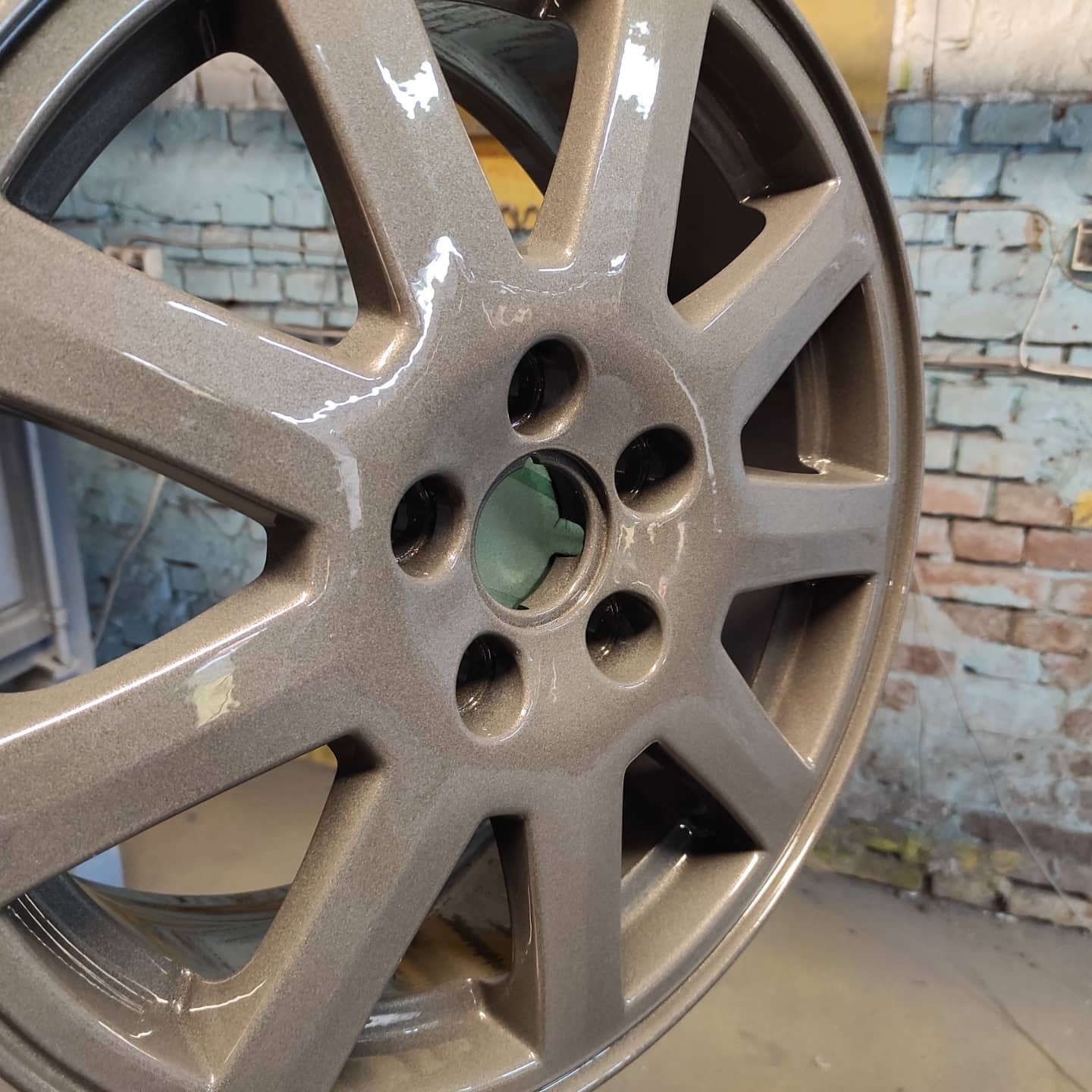 Pomalowana na szaro felga aluminiowa, widoczna na tle ściany z cegieł, z zielonym wypełnieniem w centralnym otworze.