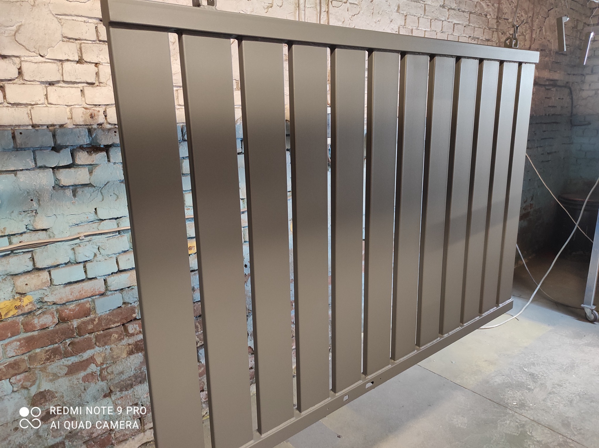 Świeżo pomalowany proszkowo szary metalowy płot lamelowy, zawieszony w warsztacie na tle ceglanej ściany z widocznymi śladami farby i instalacji elektrycznej.