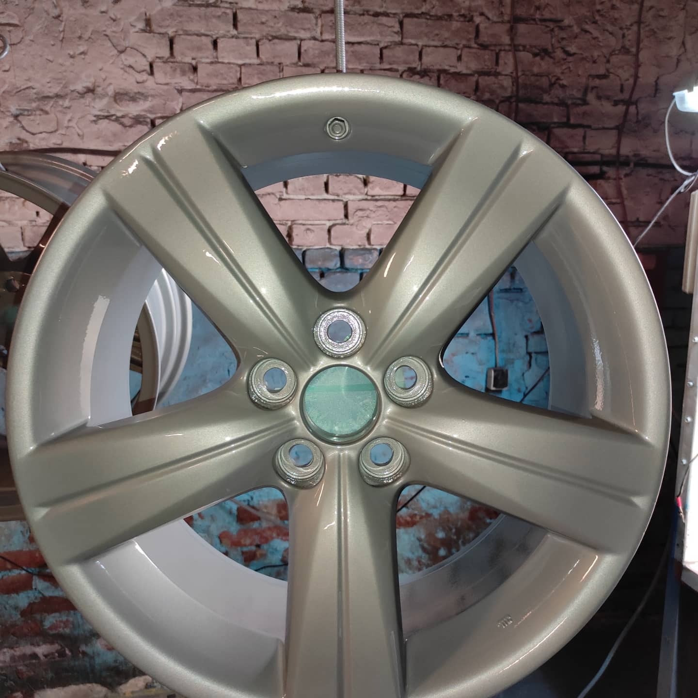 Pomalowana na złoty kolor felga aluminiowa zawieszona na lince, z widocznym efektem gładkiej powierzchni i odbiciami światła, na tle ceglanej ściany warsztatu.