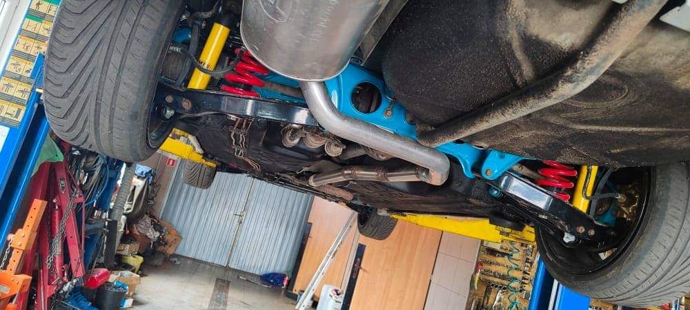 Widok podwozia samochodu na podnośniku, z widocznymi elementami zawieszenia pomalowanymi na niebiesko, żółte amortyzatory i czerwone sprężyny. Układ wydechowy w kolorze srebrnym. Widoczne narzędzia...