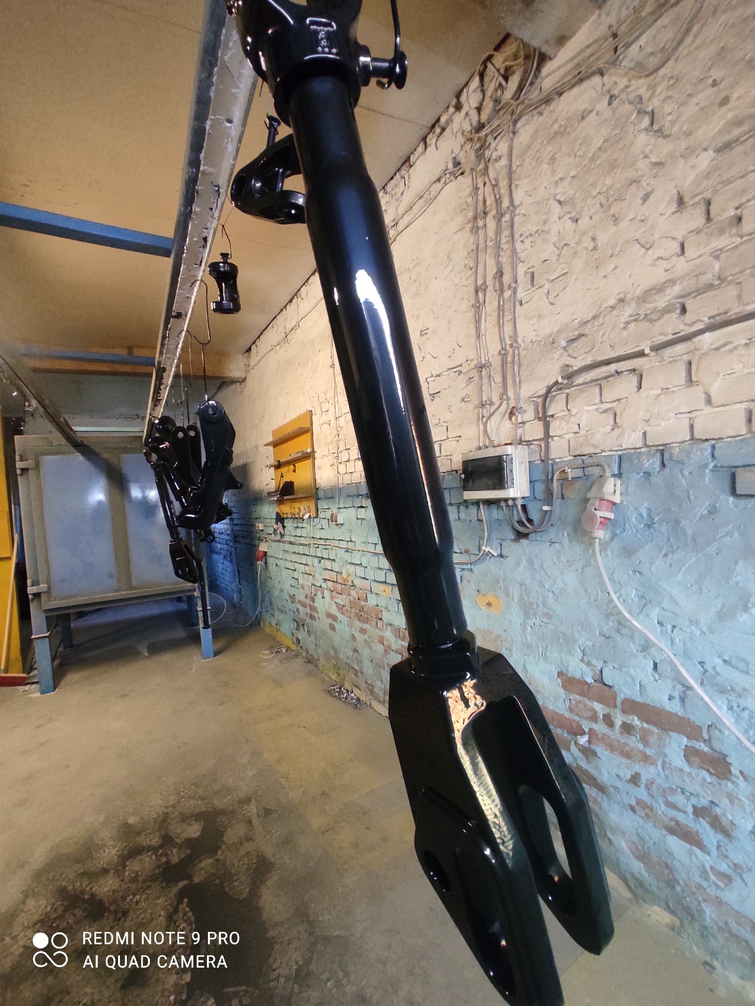 Części metalowe zawieszone na haku, pokryte świeżą, błyszczącą czarną farbą, prawdopodobnie po procesie malowania proszkowego, w tle ceglana ściana z instalacją elektryczną.