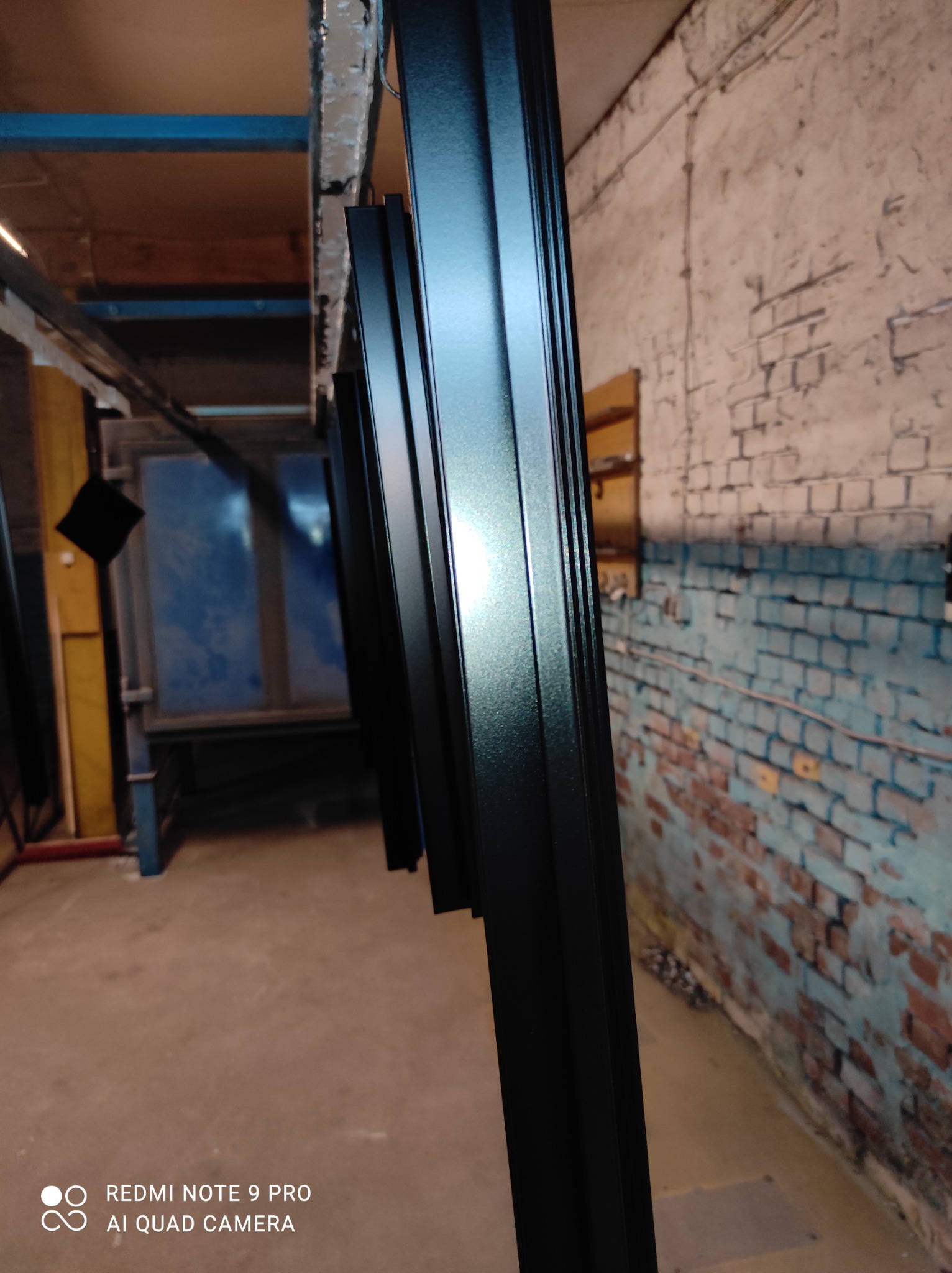 Czarne, metalowe profile zawieszone na wieszakach w hali produkcyjnej, przygotowane do dalszej obróbki lub po malowaniu, z fragmentem pieca w tle i ceglaną ścianą.