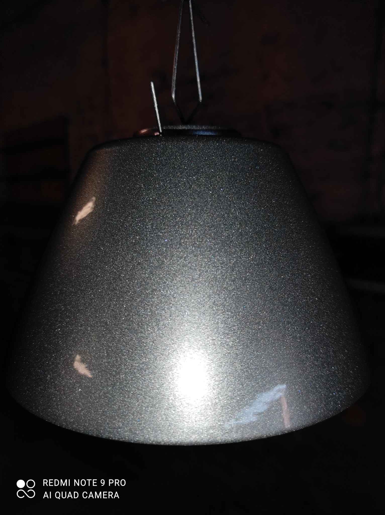 Metalowy klosz lampy w kolorze grafitowym, zawieszony na drucianym wieszaku, z widoczną fakturą po malowaniu proszkowym i drobnymi niedoskonałościami powierzchni.