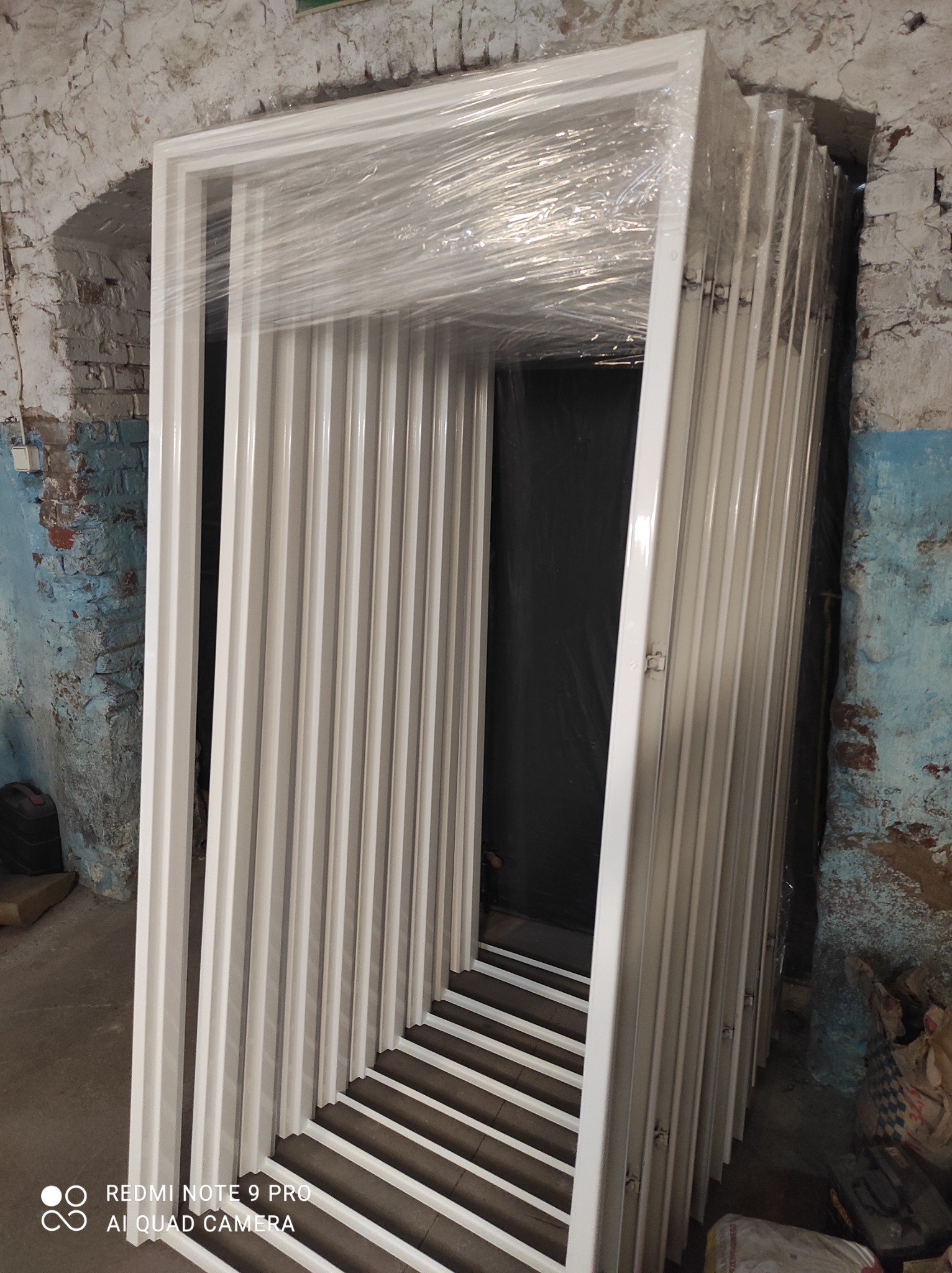 Stos białych, metalowych ościeżnic drzwiowych, zabezpieczonych folią, ustawionych pionowo w surowym wnętrzu z ceglaną ścianą.