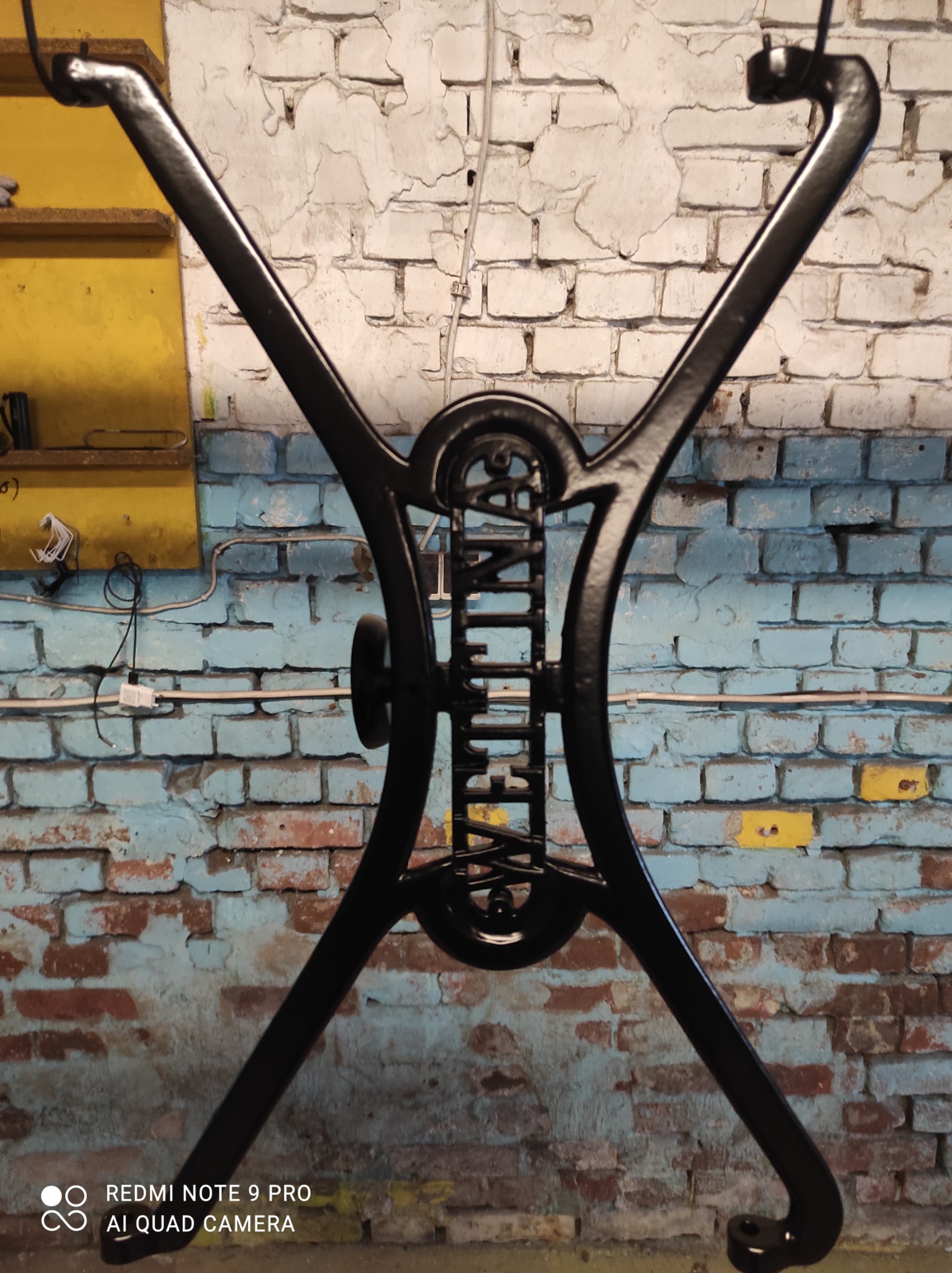 Metalowa podstawa stołu w kształcie litery X, pomalowana na czarno, z dekoracyjnymi elementami, na tle ściany z cegieł pomalowanych na biało i niebiesko, widoczne ślady użytkowania.