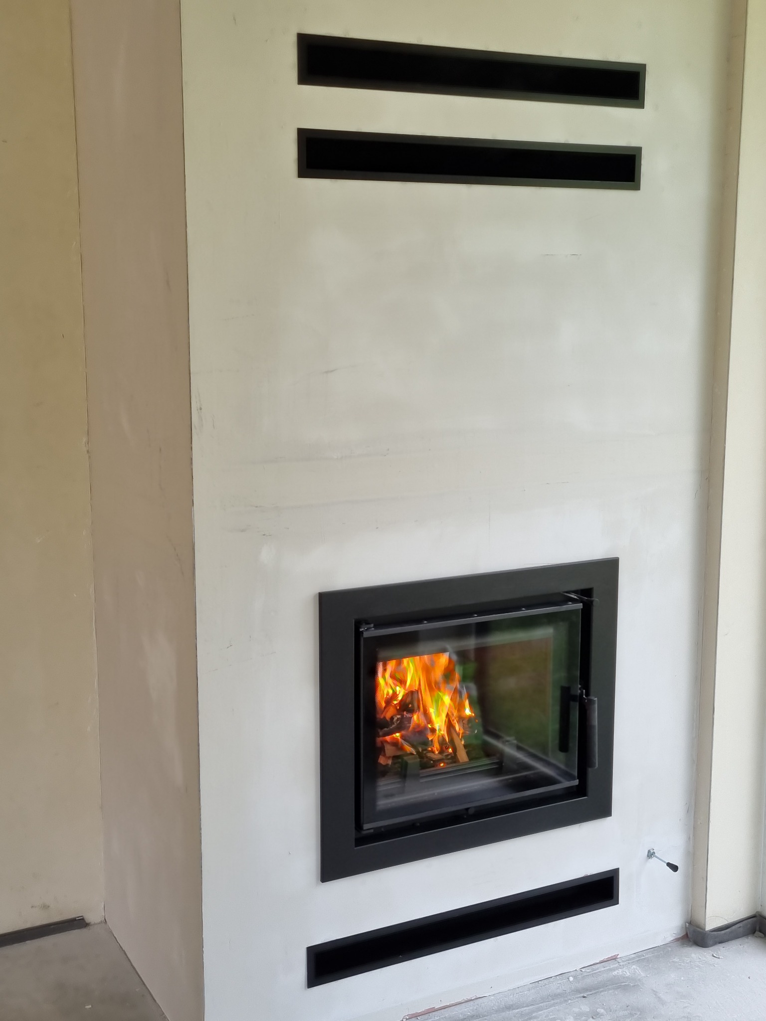 Nowoczesny wkład kominkowy z czarną ramą i płomieniami, wbudowany w białą ścianę z trzema poziomymi szczelinami wentylacyjnymi, widoczny fragment surowej ściany obok.