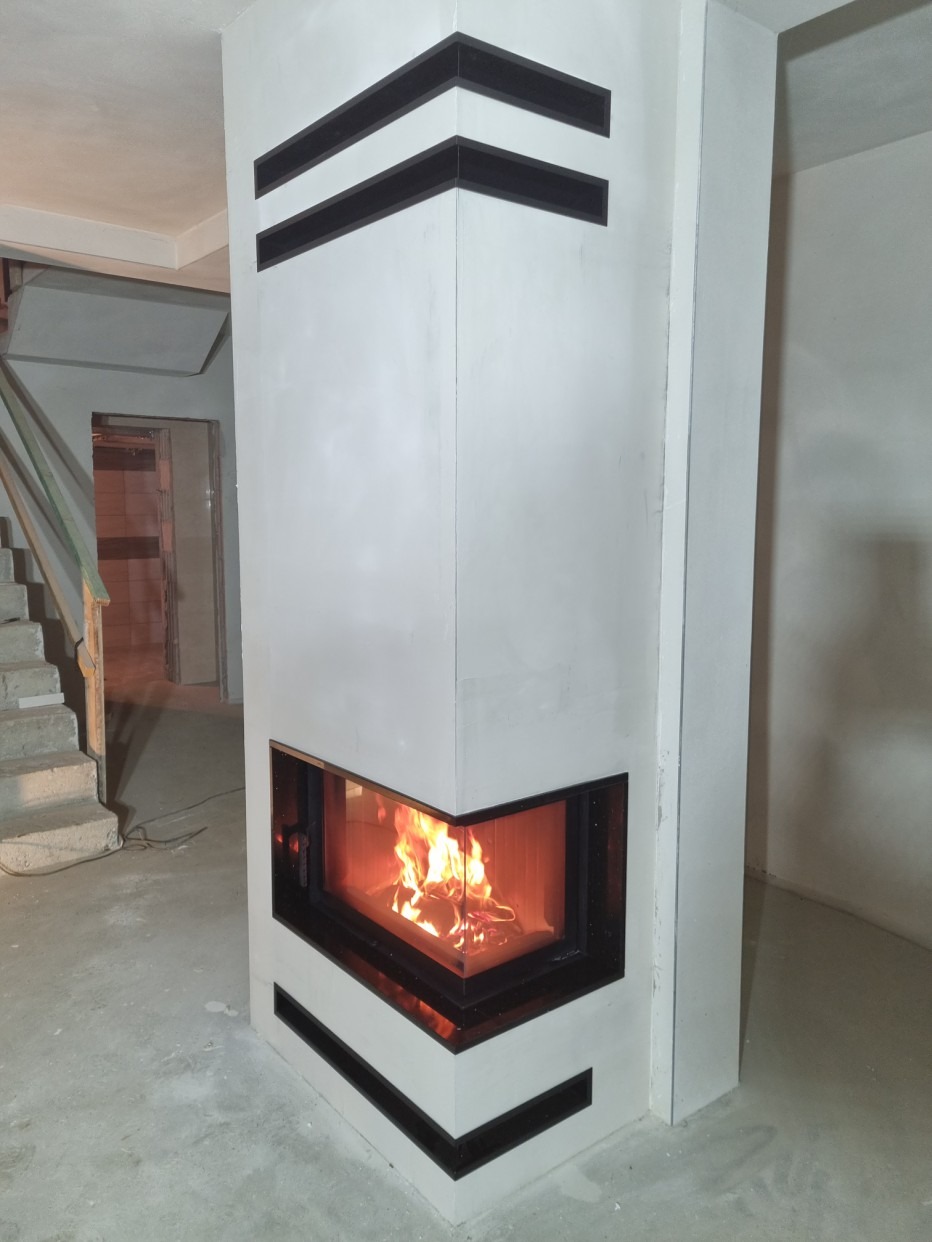 Narożny kominek z białego tynku z czarnymi, poziomymi akcentami, widoczne płomienie, surowy stan wykończenia podłogi i schodów w tle.