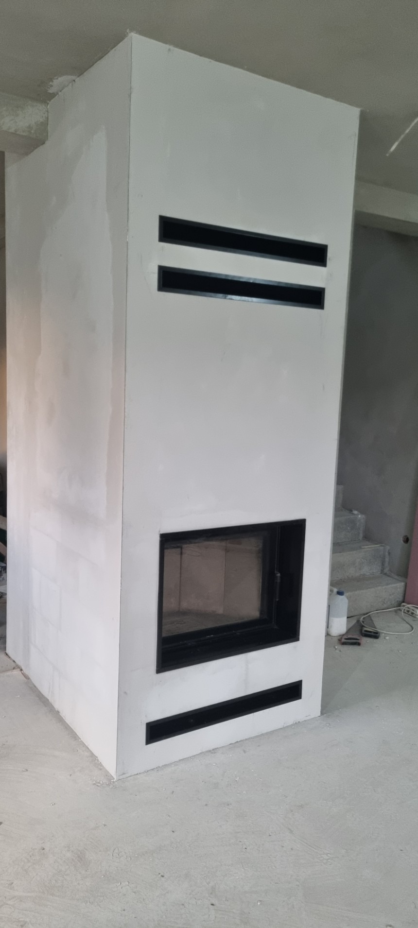 Surowy, biały korpus wkładu kominkowego z czarnymi szczelinami wentylacyjnymi, w trakcie wykończenia, widoczne ślady gładzi szpachlowej na ścianach i podłodze.
