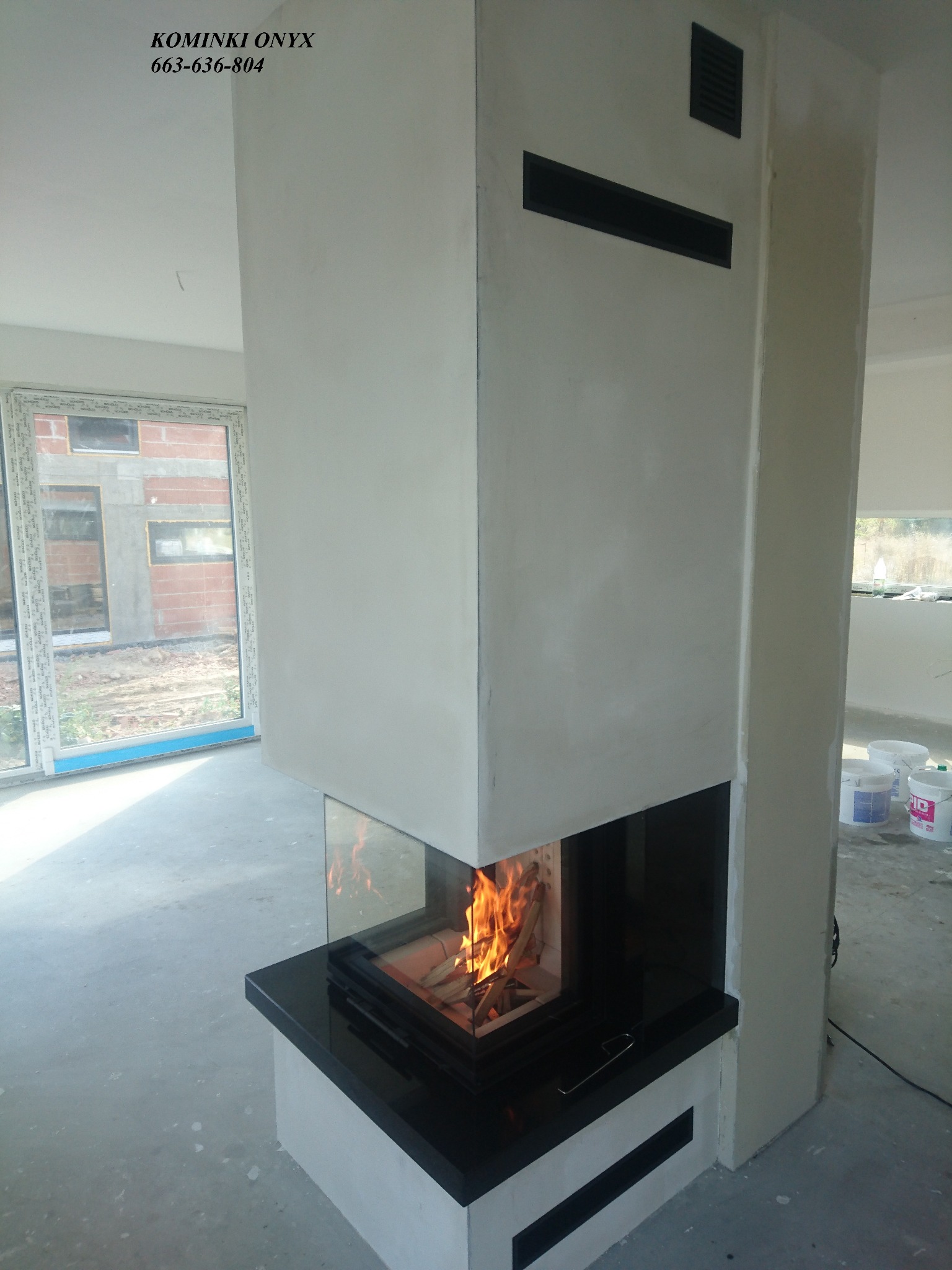 Nowoczesny kominek narożny z szybą, palenisko z płomieniami, czarny blat, biała obudowa z czarnymi wlotami powietrza, surowy stan wykończenia wnętrza z widocznym oknem i wiadrami z farbą.