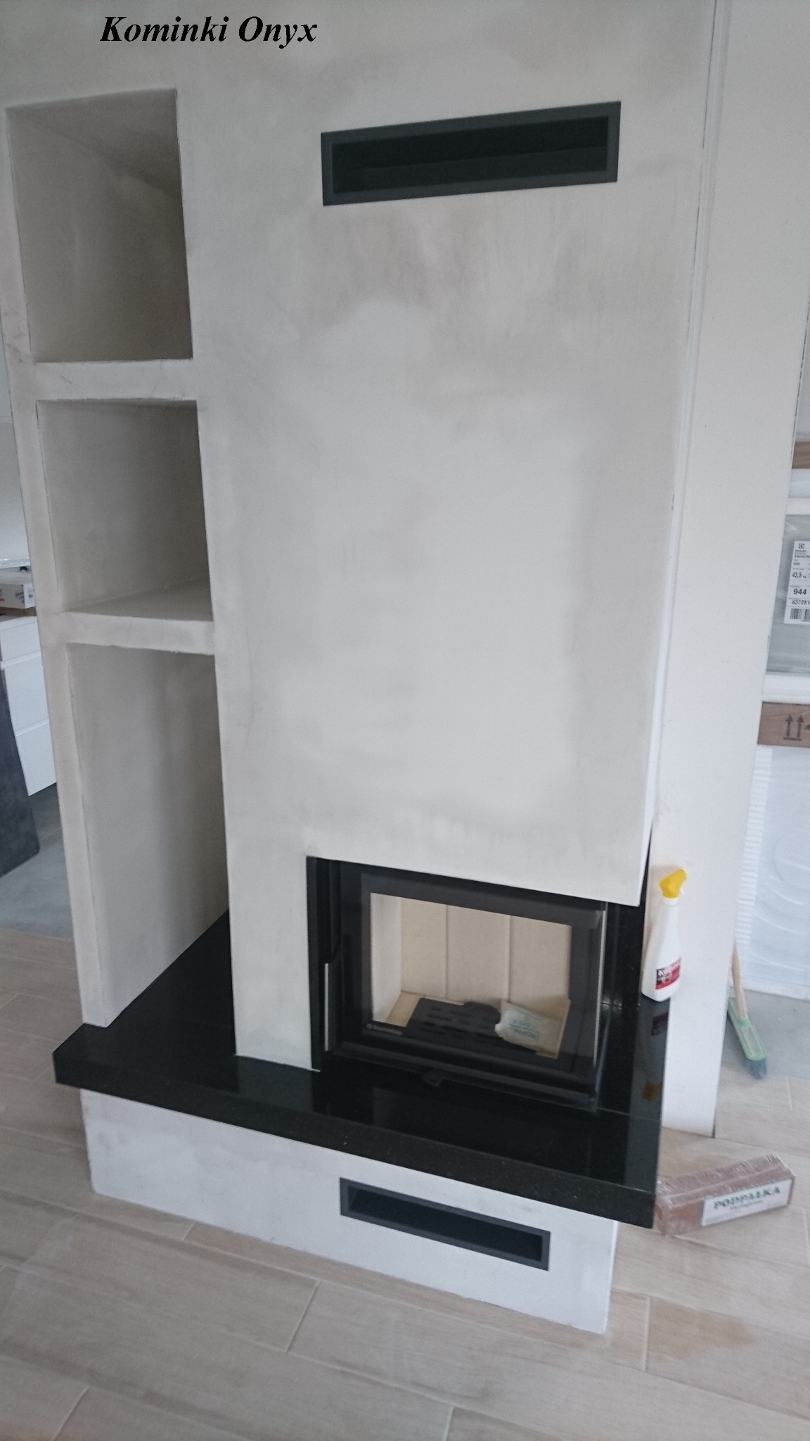 Nowoczesny, minimalistyczny piec z obudową w odcieniach bieli i czerni, wbudowany w ścianę z półkami po lewej stronie, widoczny fragment podłogi z płytek.