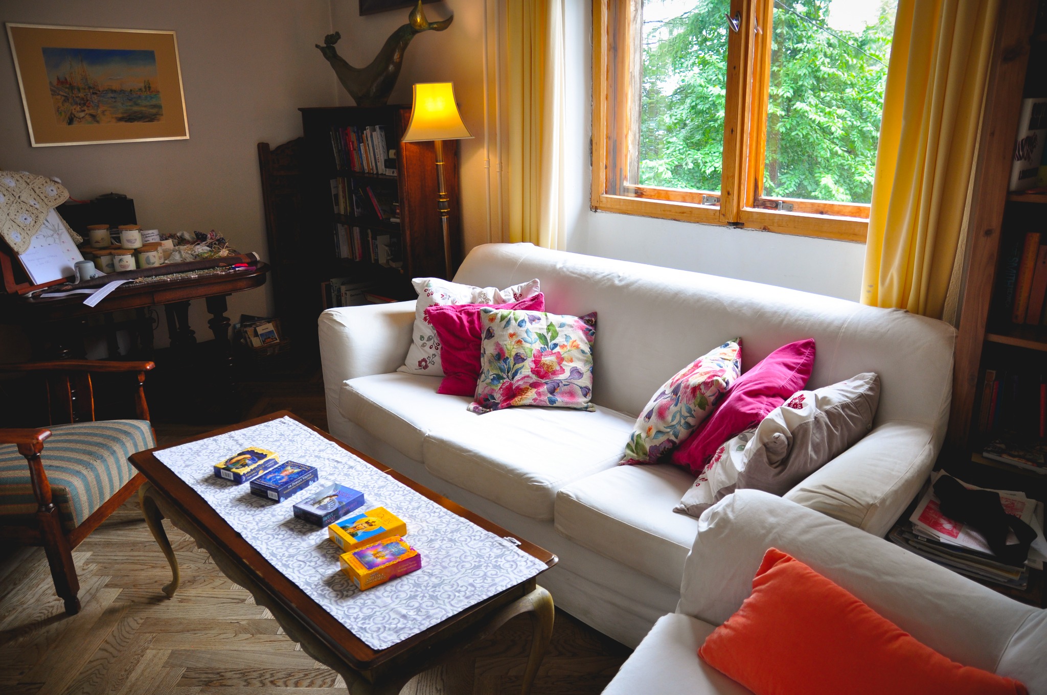 Przytulne wnętrze pokoju dziennego z białym narożnikiem, kolorowymi poduszkami i drewnianym stolikiem kawowym z rozłożonymi kartami do wróżenia, obok okna z widokiem na zieleń.