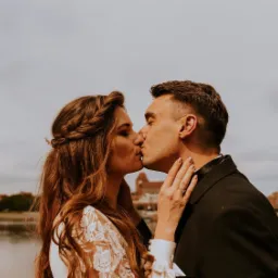 Pocałunek pary młodej na tle rzeki i zabudowań miejskich, panna młoda z warkoczem i ozdobą we włosach, widoczny pierścionek zaręczynowy.