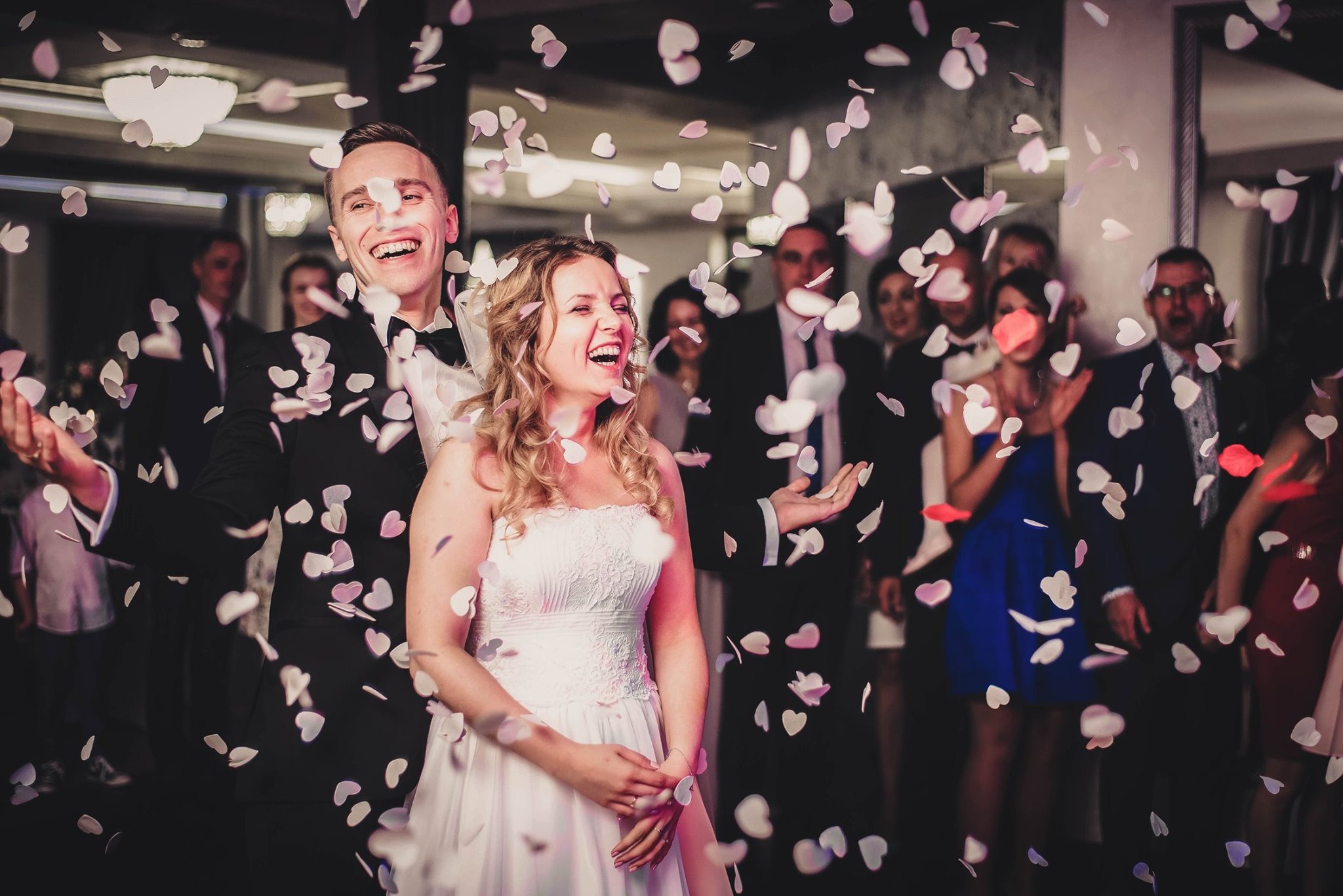 Uśmiechnięta para młoda w trakcie ceremonii zaślubin, obsypywana konfetti w kształcie serc, w tle goście weselni.