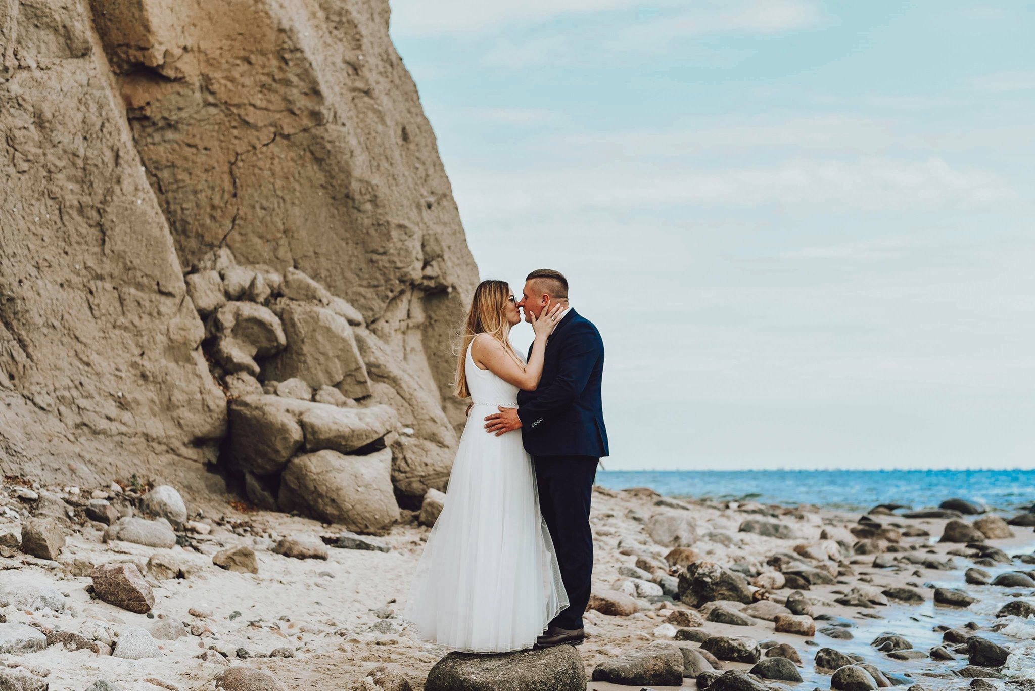 Pocałunek nowożeńców na tle klifu i morza, panna młoda w zwiewnej sukni, pan młody w granatowym garniturze, stoją na kamieniu.