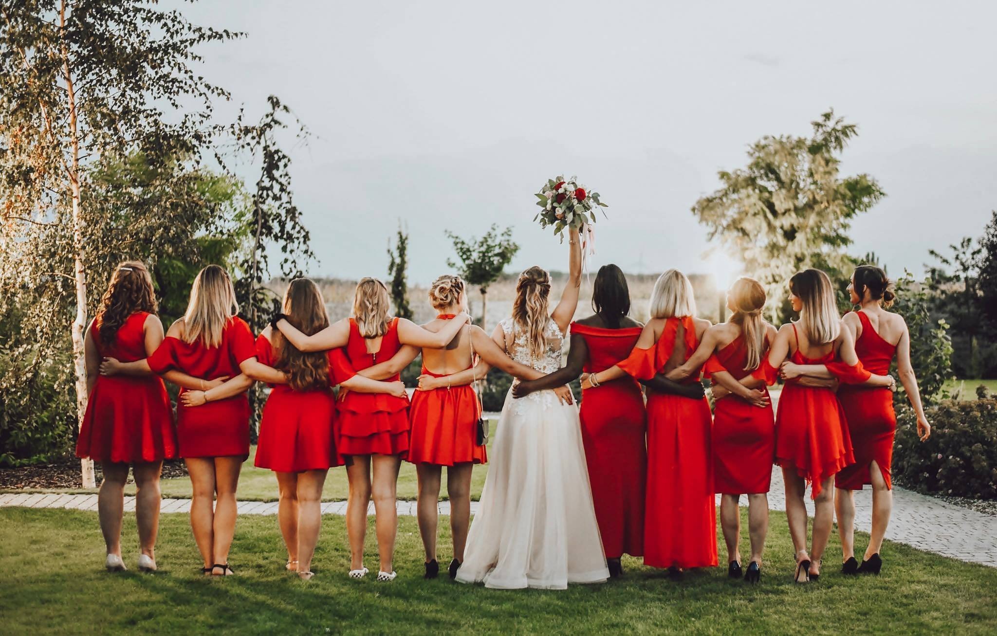 Panna młoda w białej sukni z podniesionym bukietem otoczona druhnami w czerwonych sukienkach, stoją tyłem na trawniku w plenerze.