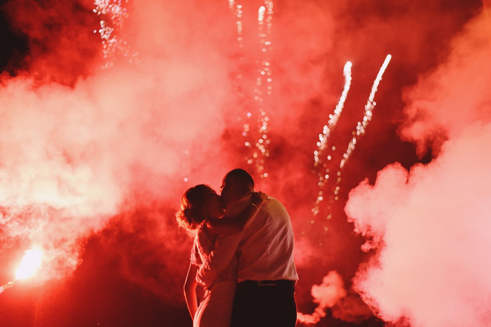 Para młoda w objęciach, pocałunek na tle czerwonych rac i fajerwerków, wieczorowa atmosfera.