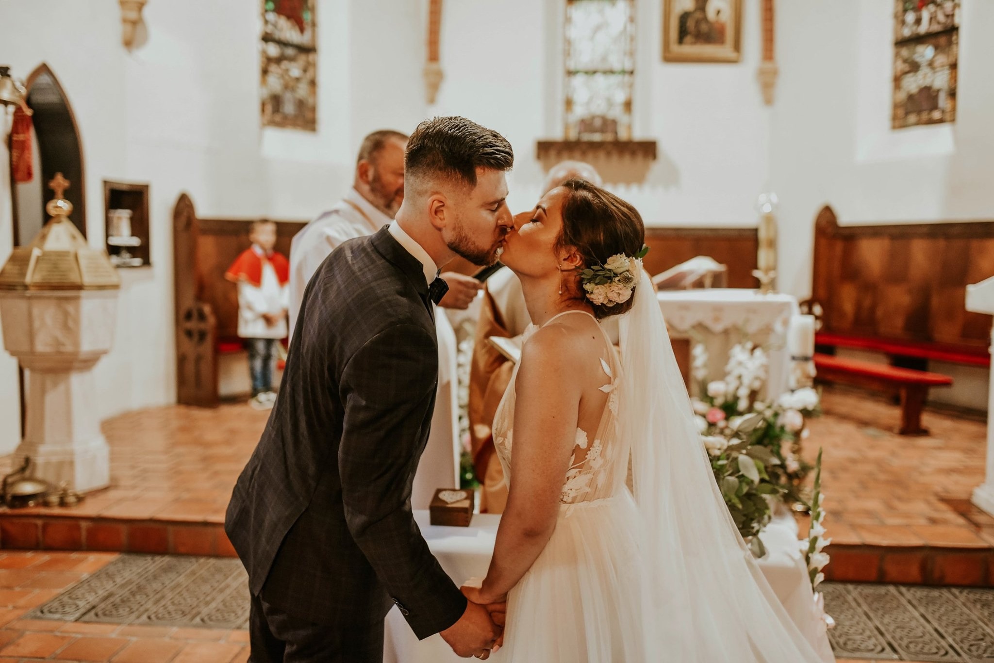 Pocałunek nowożeńców w kościele, panna młoda w sukni z aplikacjami kwiatowymi i welonem, pan młody w ciemnym garniturze z muchą, w tle ołtarz i duchowny.