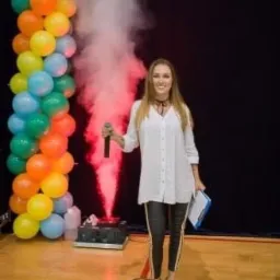 Uśmiechnięta prezenterka z mikrofonem na scenie z balonami, wytwornicą dymu i oświetleniem.