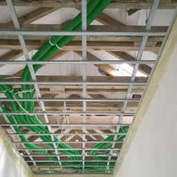 Konstrukcja metalowa sufitu podwieszanego z widocznymi drewnianymi elementami więźby dachowej oraz instalacją elektryczną w zielonych peszlach, widok z dołu.