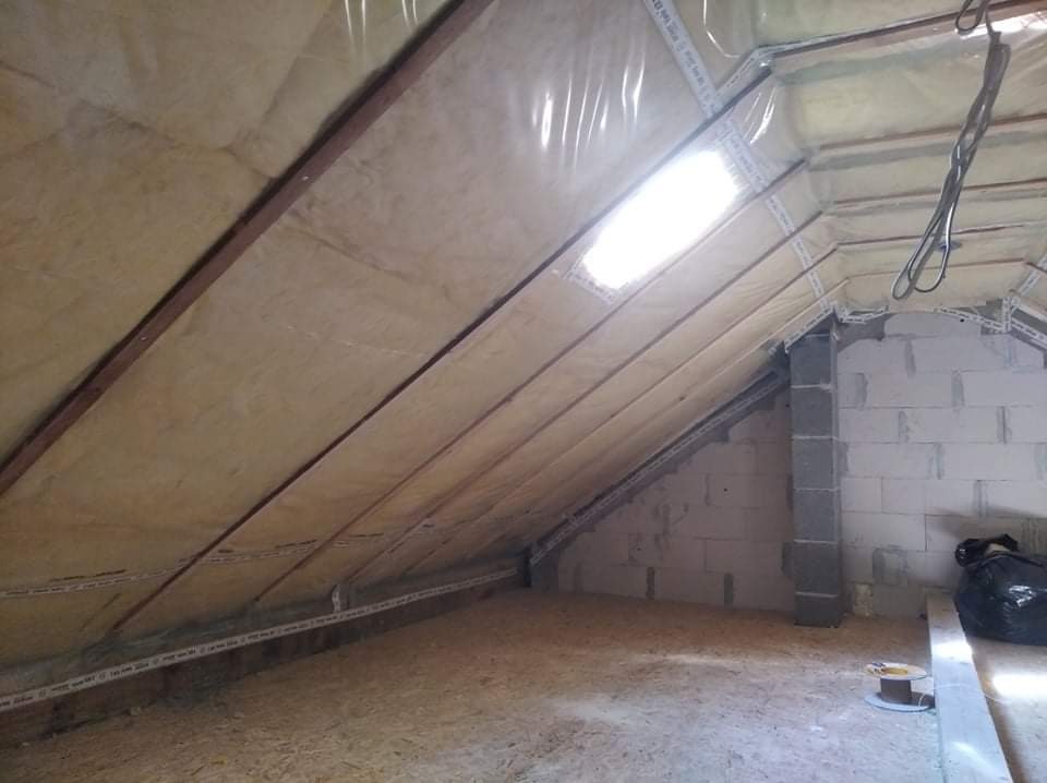 Ocieplone wełną mineralną poddasze z widocznymi krokwiami, zabezpieczone folią, z lukarną w dachu i fragmentem ściany z bloczków betonowych w tle.