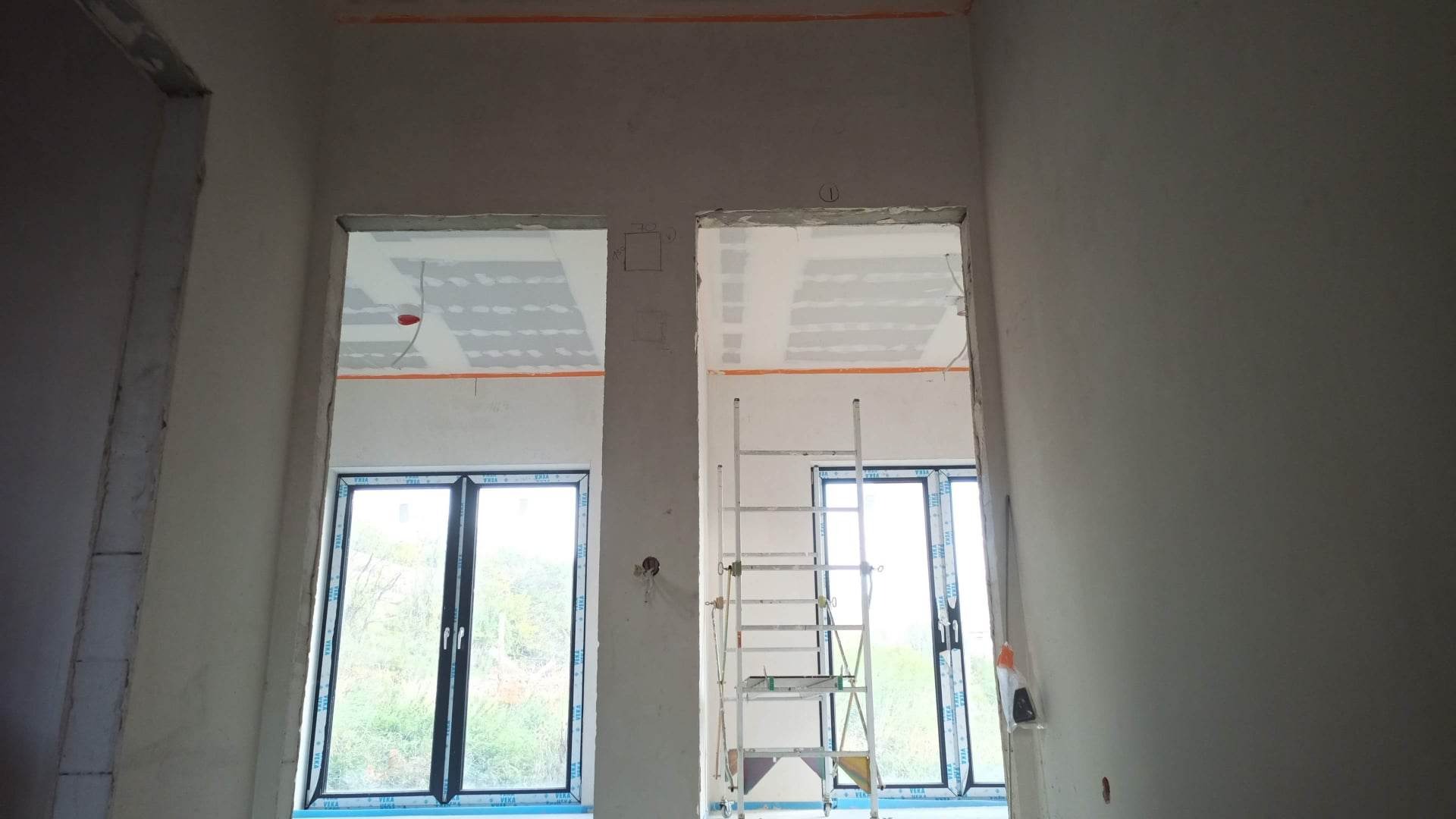 Wnętrze w trakcie prac remontowych: surowe ściany po szpachlowaniu, widoczne okna z ramami z tworzywa sztucznego, rusztowanie aluminiowe ustawione przy oknie, przygotowany sufit z płytami. Ściany...