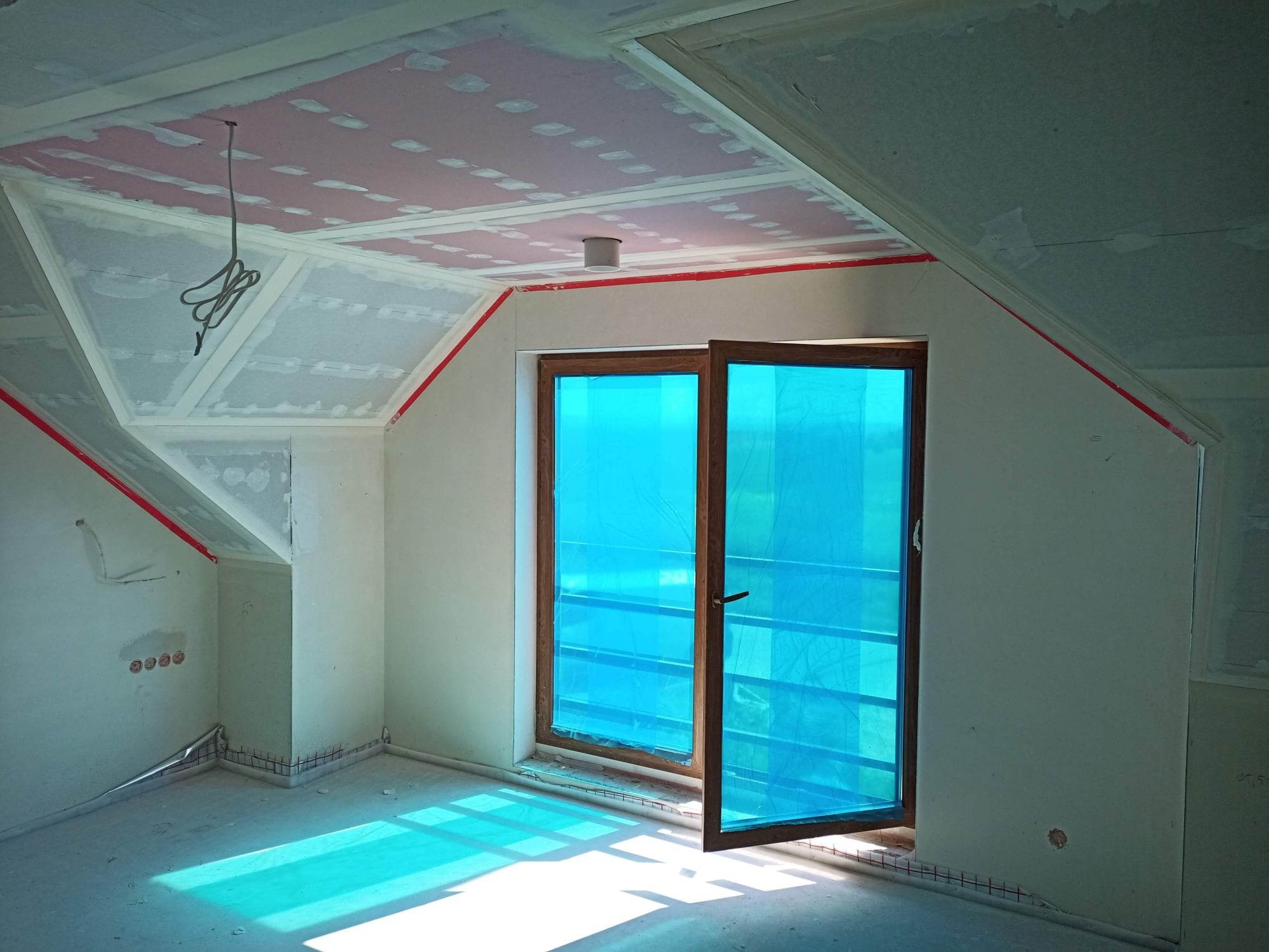 Poddasze w trakcie wykańczania: widoczne płyty kartonowo-gipsowe na skośnym suficie, zabezpieczone okno folią ochronną, ściany przygotowane do dalszych prac wykończeniowych, taśma ochronna przy...