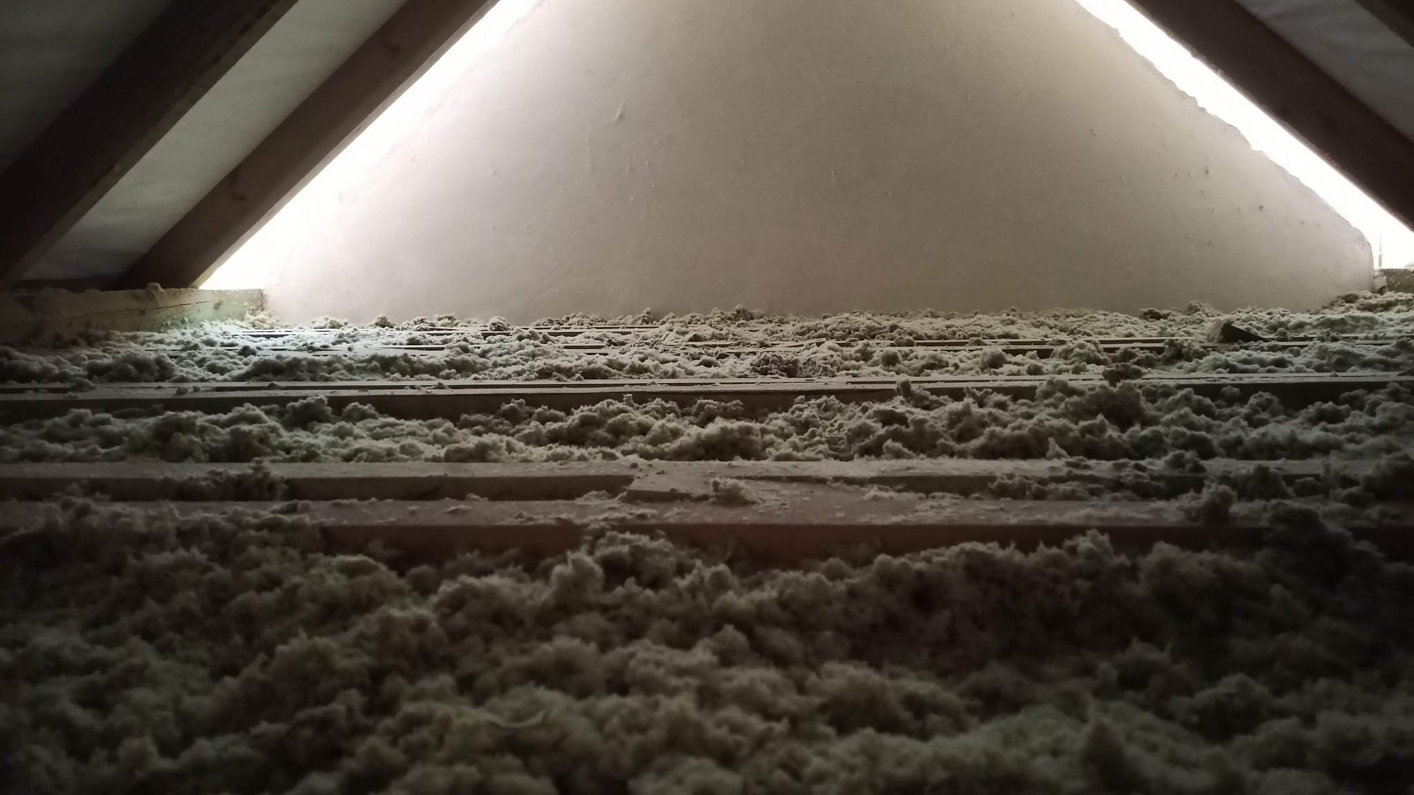 Poddasze z drewnianą konstrukcją wypełnione wełną mineralną, widoczne luki w izolacji i fragment ściany szczytowej z przebijającym światłem.