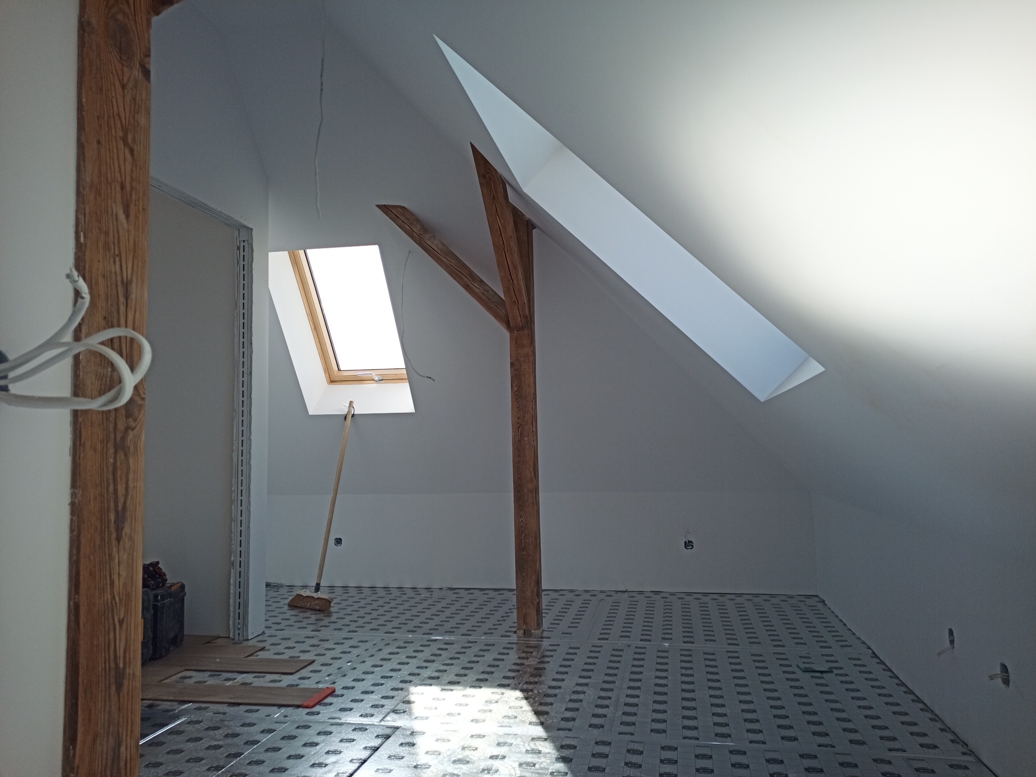 Poddasze w trakcie prac wykończeniowych: widoczne drewniane słupy konstrukcyjne, okno dachowe, oraz podłoże przygotowane pod dalsze prace, z widoczną izolacją podłogową i pozostawionymi narzędziami.